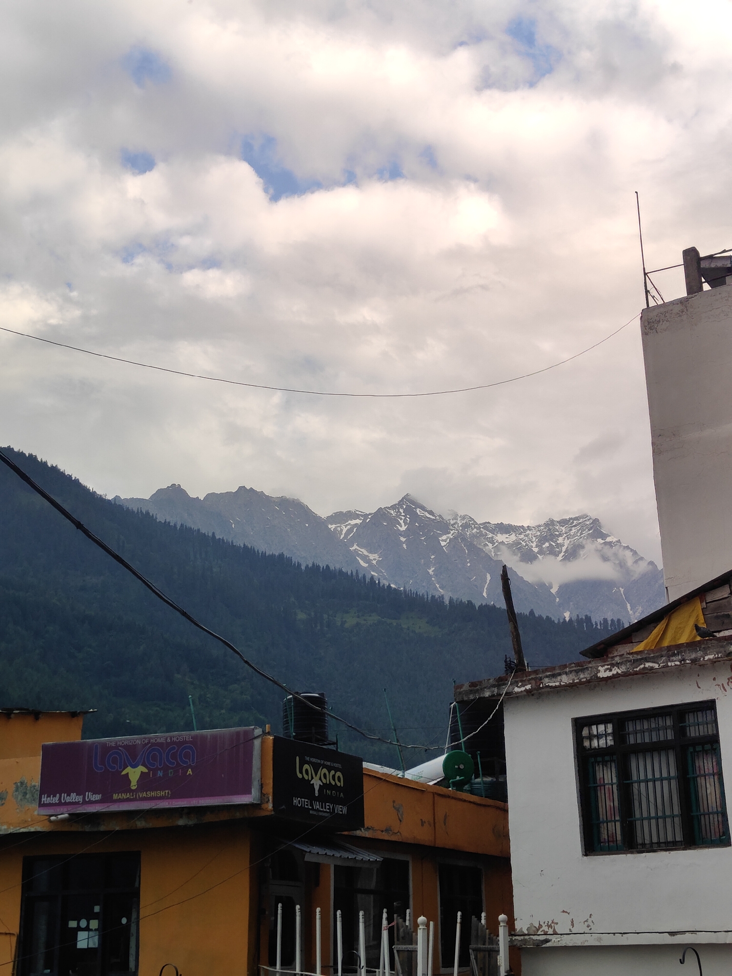 The image captures a view of the Hotel Valley View in Vashisht, India, with the majestic Himalayas in the background. The sky is partly cloudy, with scattered white clouds against a pale blue backdrop. In the foreground, two buildings are visible. The left building, painted yellow, features a prominent sign for "Lavaca India Hotel Valley View Manali (Vashisht)". The sign is rectangular with a purple border and displays the "Lavaca" logo, which appears to be a stylized cow. Below this sign, a window is visible.
To the right of the yellow building is a white structure with a dark-framed window that has a metal grille. The roofline of the white building shows some wear and tear, and a yellow tarp is visible draped over a section of the roof. A black water tank sits atop the roof of the yellow building.
Beyond the buildings, a dense, dark green forest covers the lower slopes of the mountains. The upper reaches of the mountains are snow-capped and jagged, with some clouds clinging to the peaks. Several power lines crisscross the scene, stretching horizontally across the image. The overall impression is a blend of urban infrastructure with the raw beauty of the natural landscape.