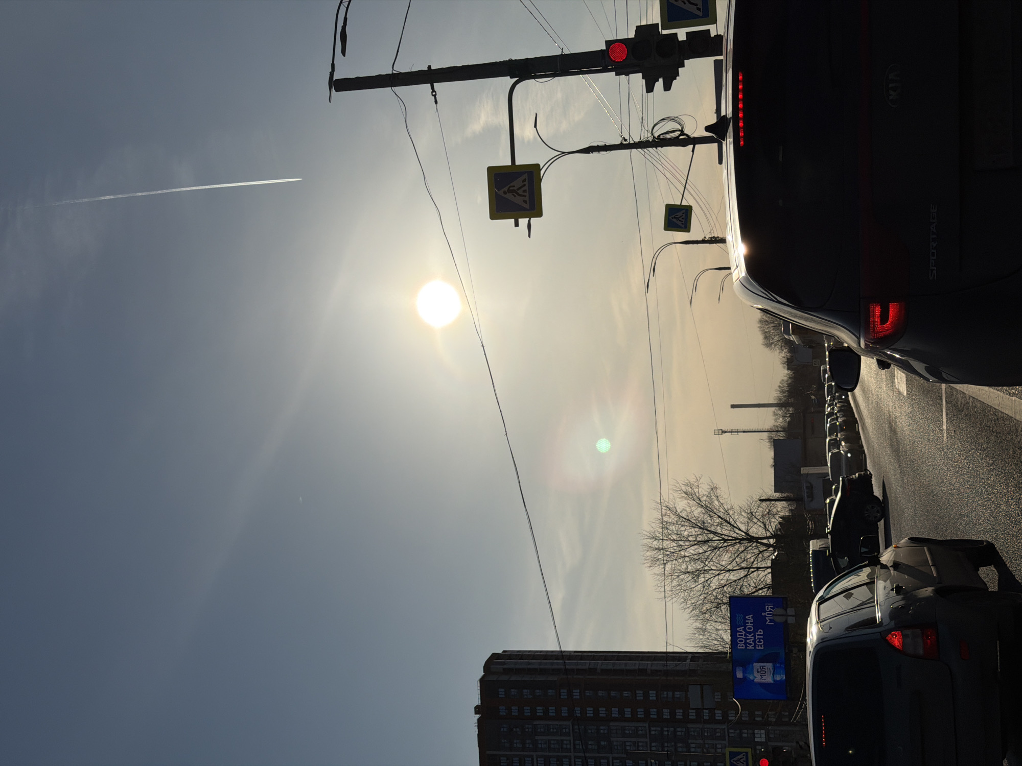 The image shows a street scene in what appears to be a city in Russia. A large apartment building is visible in the lower-left of the image. A traffic light is on red. Several cars are stopped in traffic, including a Kia Sportage immediately to the right. There is a contrail in the upper-left of the image. An advertisement is displayed on a billboard to the bottom right. The sun is high in the sky. The image is possibly taken in Izhevsk, Russia, as the billboard appears to be in Russian.