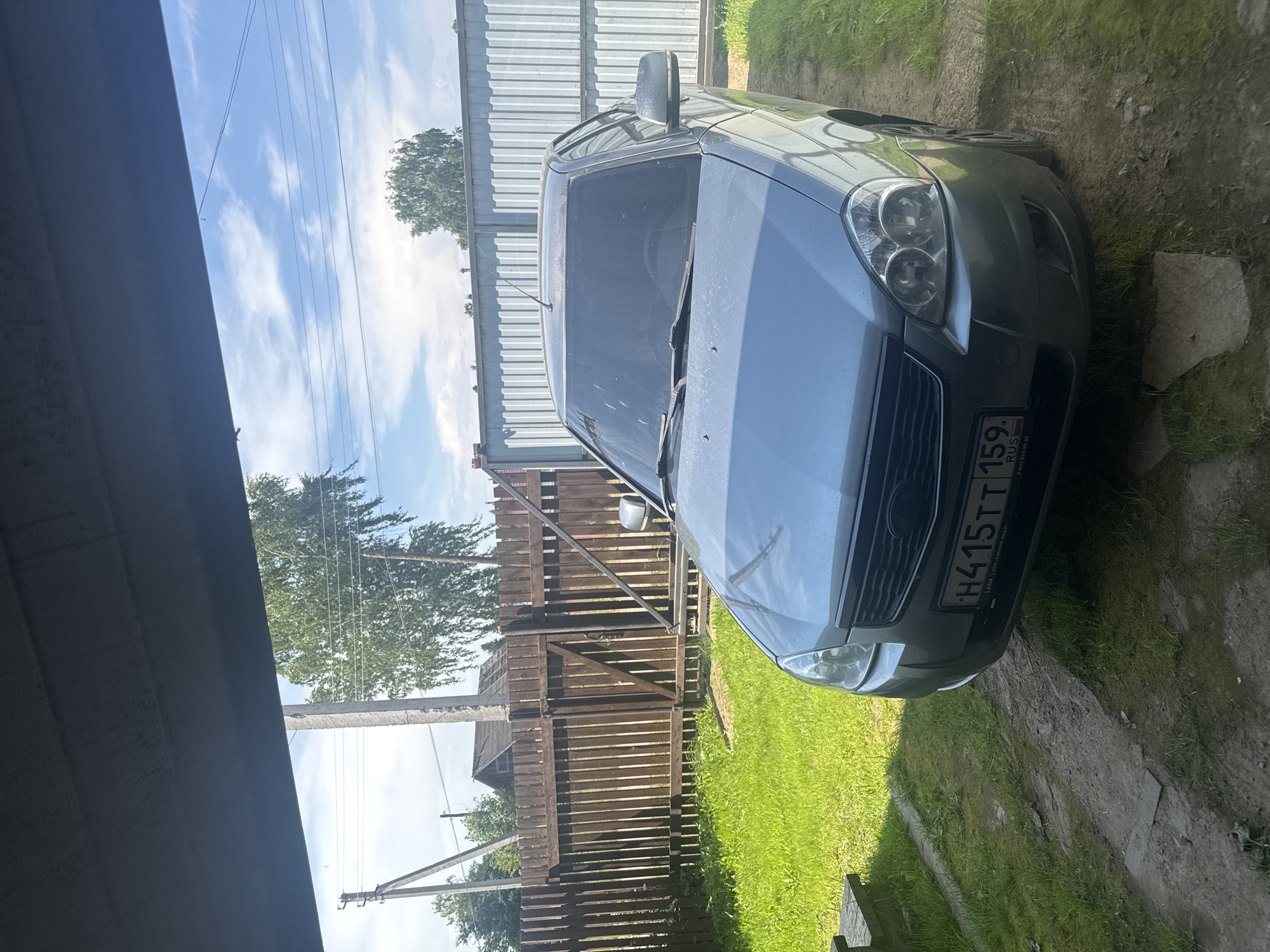 In Borodulino, Russia, a silver Lada sedan is parked in an outdoor area. The car is positioned facing forward, partially under a dark overhang that obscures the top of the frame. The vehicle's front license plate reads "H415TT 159 RUS". To the left of the car is a patch of green grass and a wooden bench. Behind the car, a wooden fence separates the yard from a grassy area and a corrugated metal fence. Trees and utility poles are visible in the background under a bright, partly cloudy sky. The ground in front of the car appears to be a dirt path.