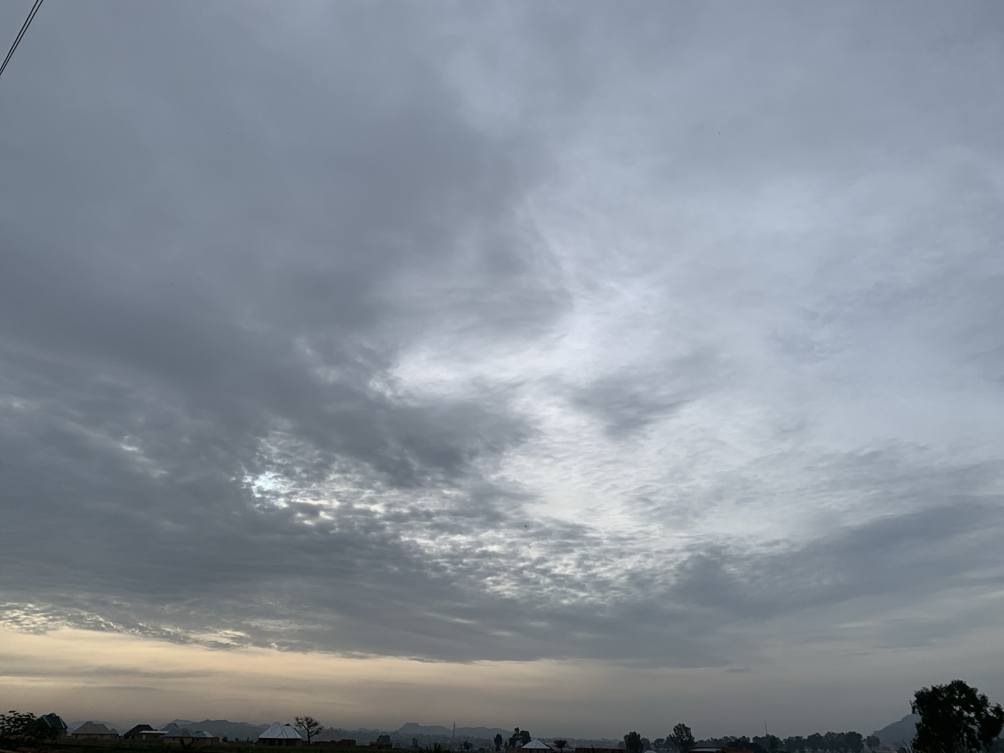 An aerial, wide-angle shot captures a cloudy sky over a rural landscape in Kuru Karama, Nigeria. The sky is filled with a mix of gray and white clouds, with patches of sunlight breaking through, creating a diffused light. Below, a village is visible, characterized by scattered houses with corrugated iron roofs. The terrain appears flat, with distant hills forming a hazy backdrop. Vegetation, including trees and shrubs, is present throughout the scene. The overall atmosphere is serene, with no apparent human activity or prominent actions captured.