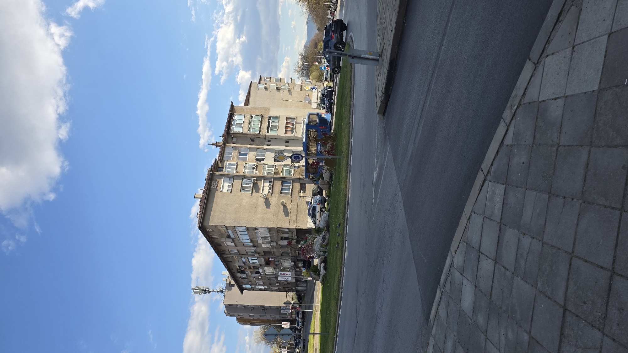 The image depicts a street scene in Blagoevgrad, Bulgaria. A multi-story residential building dominates the left side of the frame, with a blue sky and scattered clouds in the background. The building is constructed of concrete. On the right, there is a paved road and a tiled sidewalk. Several cars are parked along the street. The scene is generally sunny.