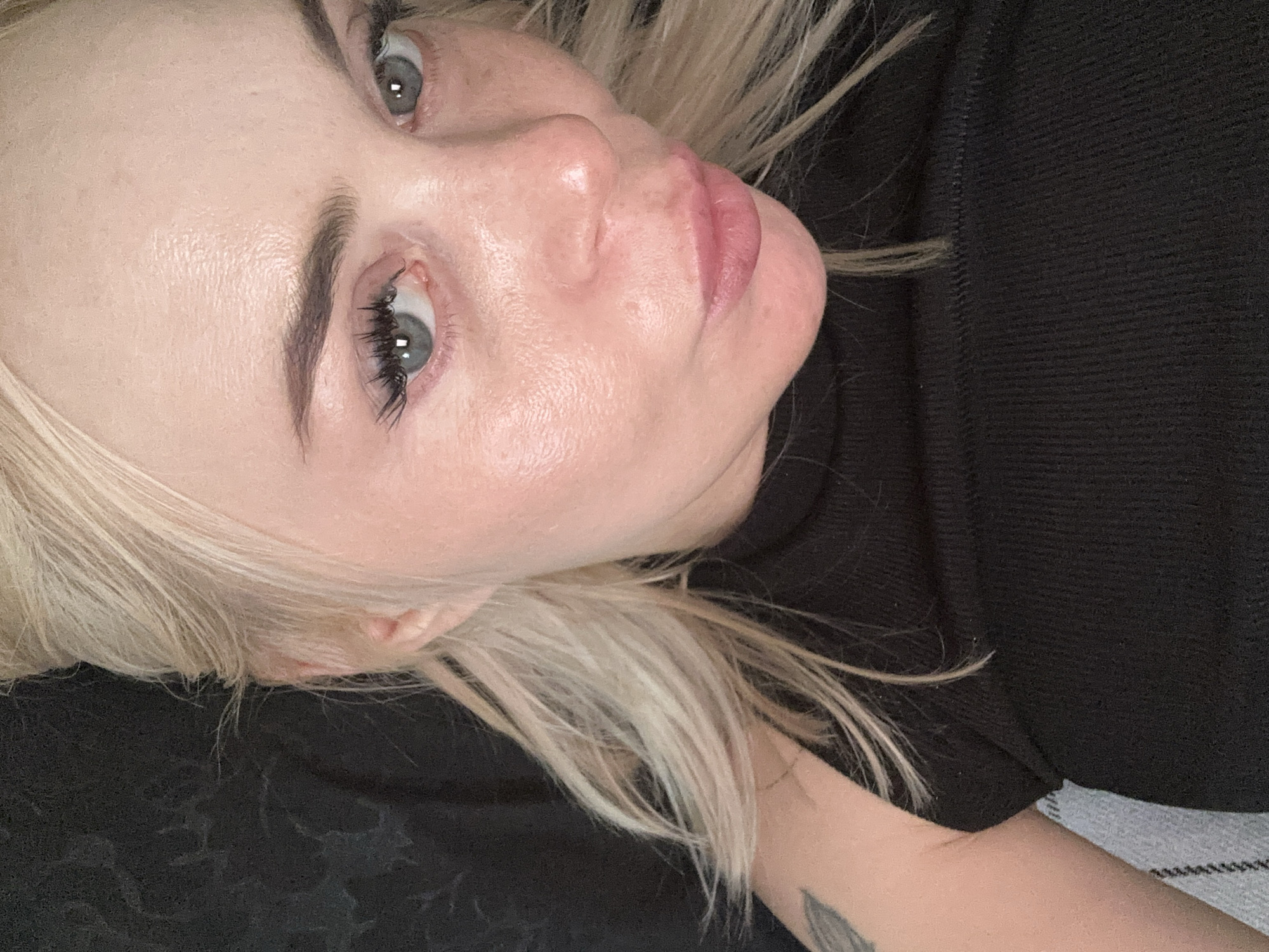 A close-up, slightly high-angle photograph captures an adult female with fair skin and platinum blonde hair, looking directly at the camera with a slight pouting expression. Her hair is styled with some strands framing her face and ears, and the bulk of it appears to be pulled back. Her blue-grey eyes are prominent, featuring defined eyebrows and long, dark eyelashes. Her skin displays natural texture, some visible pores, and a slight sheen across her forehead and nose. She wears a black, ribbed knit sleeveless top, partially revealing her left upper arm which bears a grey-toned tattoo featuring a stylized feather-like element. The background behind her is largely dark and indistinct with a subtle mottled or patterned texture, possibly fabric or a wall. To the lower left, a section of light grey and white striped fabric is visible, suggesting she is reclining or seated. The setting is in the city of Unknown, Unknown.