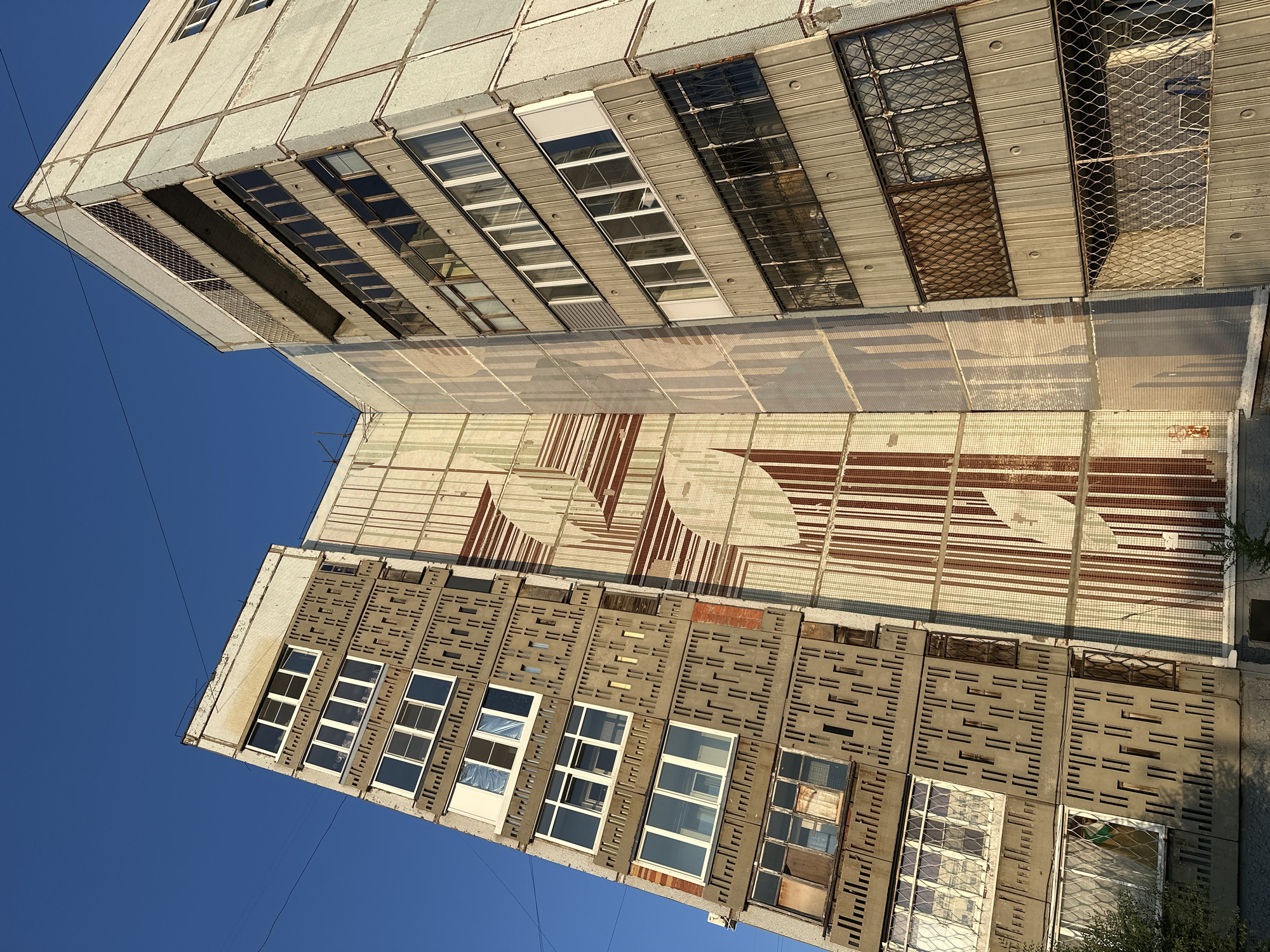 The image shows a view of a multi-story apartment building against a clear blue sky. The building appears to be constructed from concrete panels and features a patterned facade on one side, with rows of windows and balconies on the other. The building's exterior shows signs of age and wear, with some windows appearing dirty or covered. Electrical wires are visible crisscrossing the sky. The building is located in the city of Biysk, Russia.