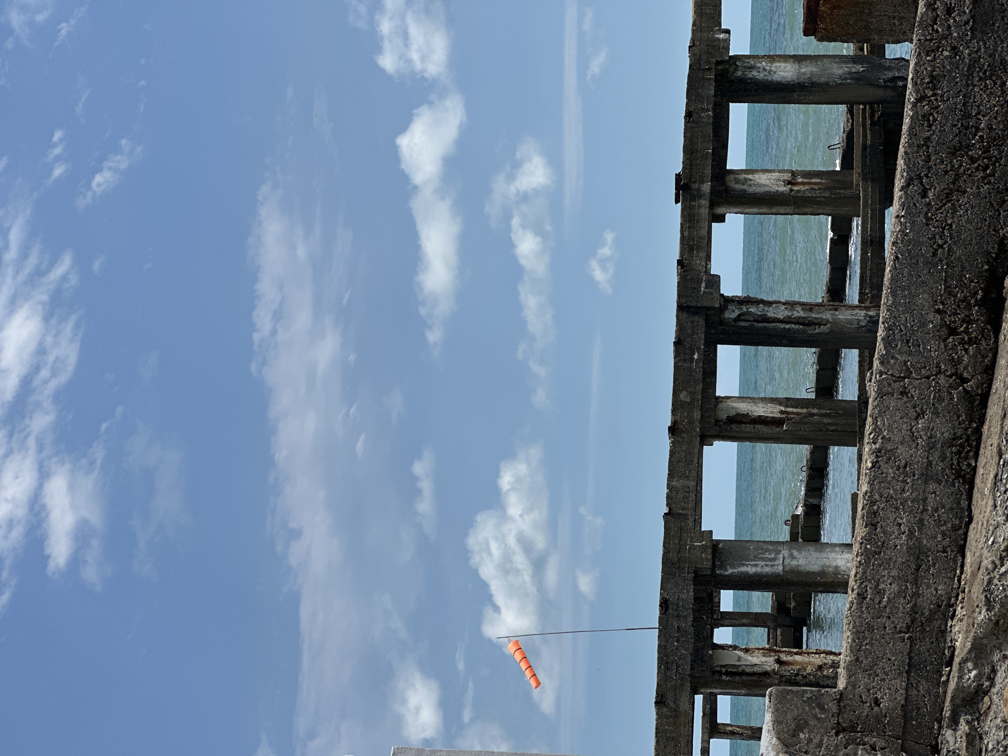 An outdoor, daytime, medium shot captures the weathered ruins of a concrete structure situated on the coast of the Black Sea in Sochi, Russia. The foreground is dominated by rough, gray concrete blocks, some appearing partially crumbled. Beyond these, a series of vertical concrete pillars support horizontal beams, forming open rectangular sections that frame the sea and sky. The ocean, a deep blue-green, is visible through these openings, with gentle waves lapping at the submerged remnants of what was likely a pier or jetty.
In the mid-ground, a thin, dark pole extends upwards, bearing an orange and white striped windsock, indicating a moderate breeze. The sky above is a clear, bright blue, dotted with scattered white cumulus clouds. The overall scene conveys a sense of decay and abandonment, with the natural elements of the sea and sky contrasting with the man-made ruins. No people are present in the image.