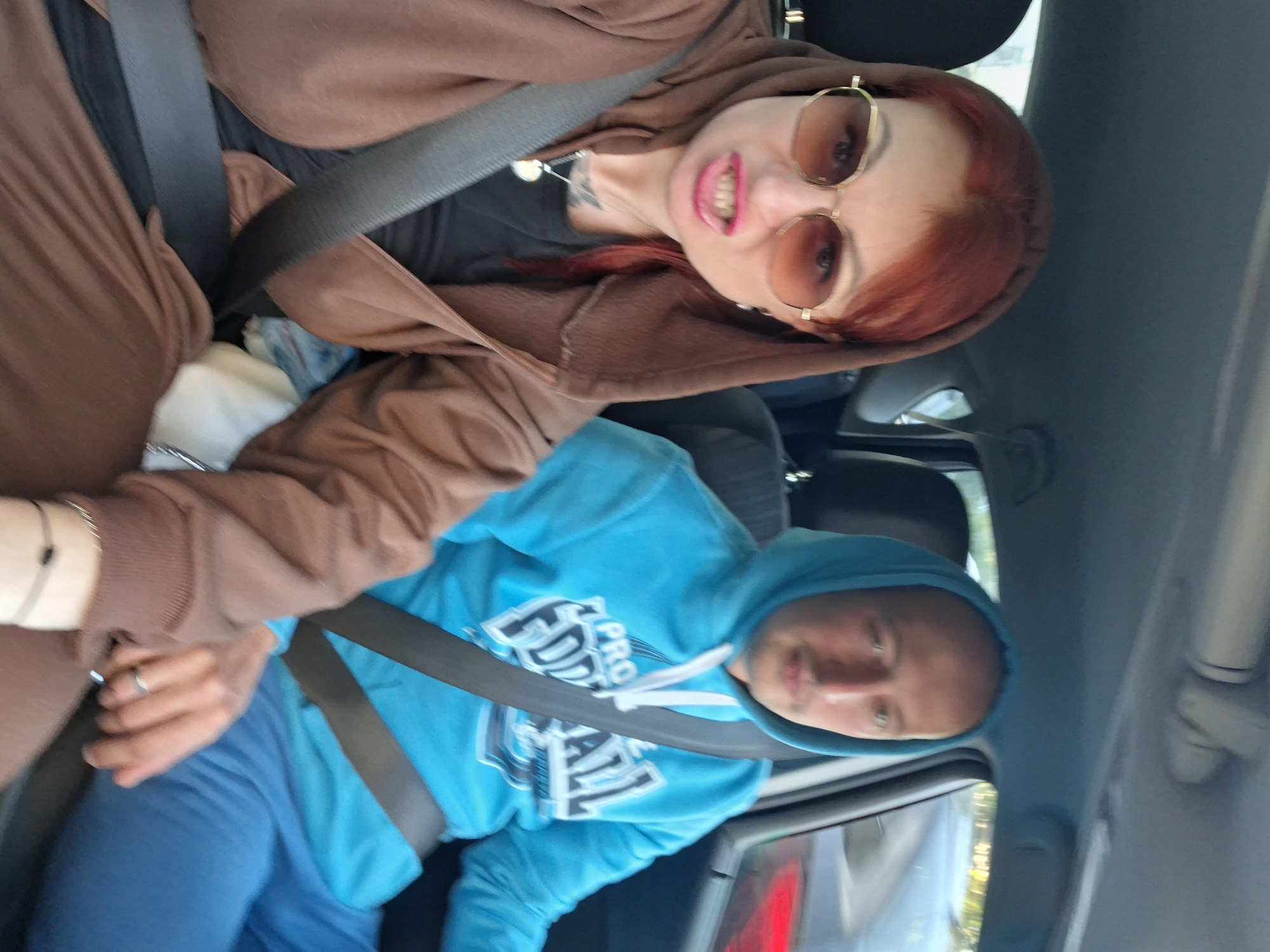Two individuals are seated inside a car. The person in the foreground, on the left, is a woman with red hair, wearing large sunglasses, a brown hooded jacket, and a black shirt. She is looking towards the camera with an open-mouthed expression. A seatbelt is visible across her chest. She is wearing a necklace with a circular pendant. In the background, on the right, a man with a shaved head is wearing a blue hooded sweatshirt and blue pants. He is also wearing a seatbelt and looking forward. The interior of the car is visible, including the dashboard and seats. The scene appears to be an urban setting, possibly in Myadzyel, Belarus, given the presence of other vehicles in the background.