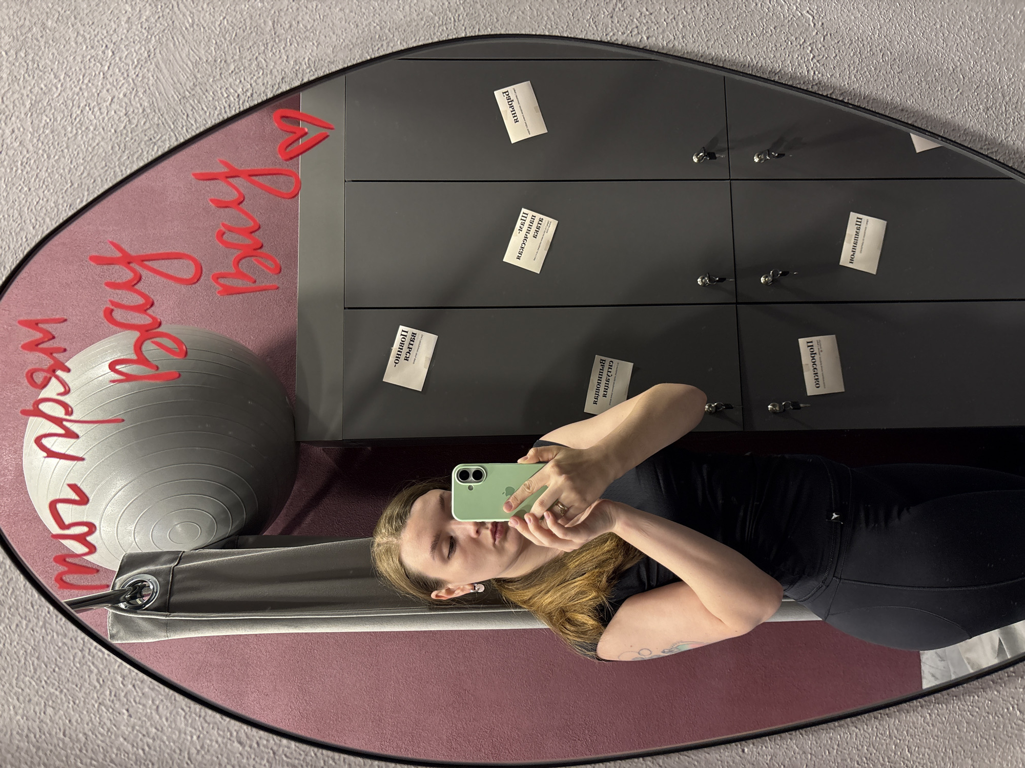 This mirror selfie, taken in Izhevsk, Russia, captures a young woman in an oval-shaped mirror, holding a light green iPhone to take the picture. She has light brown hair, wears a black athletic top and leggings, and has a subtle tattoo on her left arm. The setting appears to be a gym or fitness studio, indicated by an exercise ball visible on the left and a row of dark grey lockers behind her.
The mirror itself is decorative, featuring bold red 3D lettering that reads "ты прям вау вау!" (meaning "you're just wow wow!") along with a heart symbol. Attached to the lockers are numerous small white notes, each containing positive affirmations, reflected upside down. These affirmations, in Russian, include phrases such as "Я - идеальна" (I am ideal), "Я - прекрасна" (I am beautiful), "Я - богиня" (I am a goddess), "Я - счастлива" (I am happy), "Я - успешна" (I am successful), "Я - люблю себя" (I love myself), and "Я - источник любви" (I am a source of love), among many others, all starting with "Я" (I am). The overall scene conveys a positive, self-affirming atmosphere within a fitness environment.