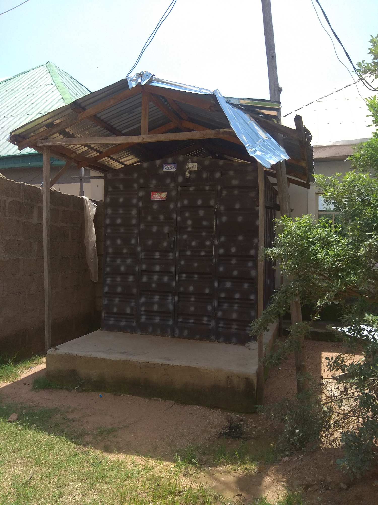 The image depicts an outdoor scene in Jalingo, Nigeria, featuring a simple, open-sided structure with a corrugated metal roof supported by wooden beams. This structure shelters a large, dark-colored metal door, possibly the entrance to a room or storage area. The metal sheeting on the door has a repeating pattern of white dots. To the left of the structure is a rough, concrete block wall. A white plastic sheet is draped over the top of this wall. The ground in front of the structure is a mixture of soil and sparse grass. To the right, a leafy green bush or small tree grows near the structure, with its branches partially obscuring the view of the building. Electrical wires are visible overhead, strung against a clear, bright sky. The overall impression is one of a utilitarian, possibly informal, building or enclosure in an outdoor setting.