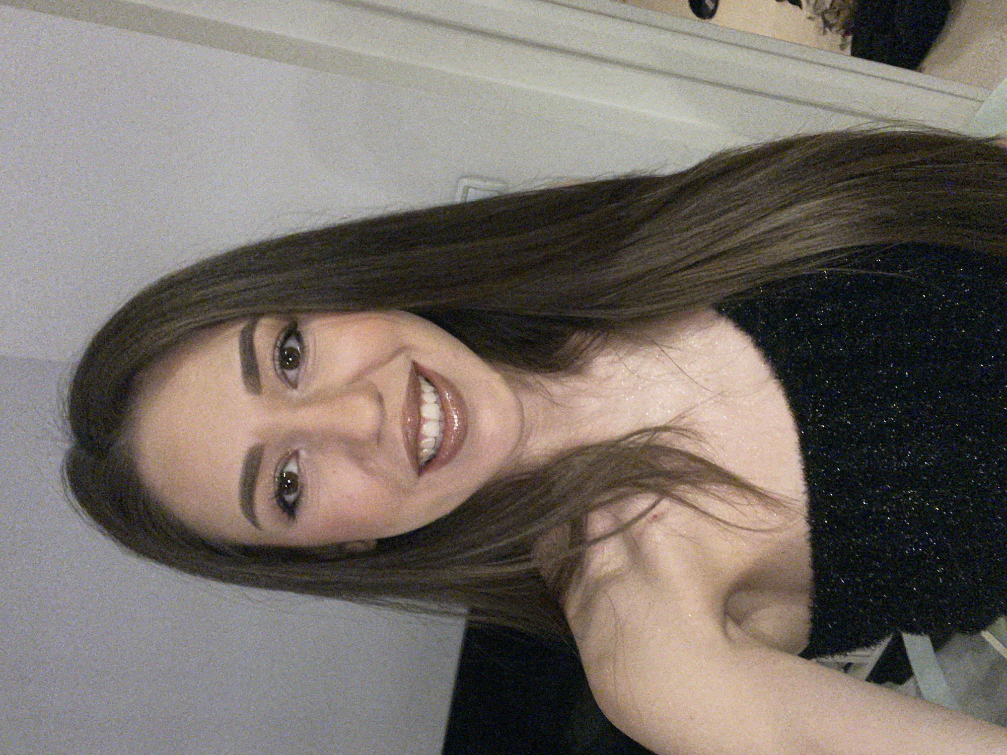 A young woman is photographed indoors. Her dark, straight hair falls past her shoulders. She is smiling, revealing white teeth, and is wearing a black sparkly top. The background is a light gray wall with a white door frame at the top. The location is unknown; there is no indication it is Sofia, Bulgaria.
