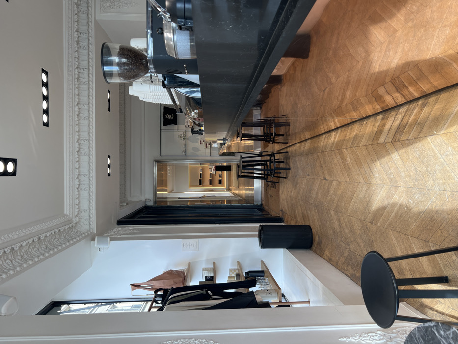 The image displays the interior of a commercial establishment located in Paris, France, functioning as a boutique or concept store with an integrated coffee bar. The perspective is from the front, looking towards the rear of the space.
On the left, a retail display area features a white wall with three tiered light wood shelves holding various merchandise, including bottles and candles. A clothing rack to the left of the shelves exhibits several dark garments and a light brown paper tote bag. A tall, black cylindrical trash receptacle stands on the floor near the wall. A tall window with black framing allows natural light into the room and offers a glimpse of an external building with an ornate railing.
The right side of the image is dominated by a long, dark counter functioning as a coffee bar. Visible equipment on the counter includes a coffee grinder filled with roasted coffee beans, stacks of white disposable cups, and various metal containers associated with coffee preparation. The lower section of the counter has a highly reflective surface, mirroring the chevron-patterned wooden floor.
The central floor area is covered with a meticulously maintained wooden floor laid in a chevron pattern, extending towards a lighted doorway at the far end. This doorway leads to a secondary room or corridor, where illuminated shelves displaying additional merchandise are visible. Three sets of black metal bar stools are arranged in front of the coffee bar, and a single black stool with a round seat is present in the immediate foreground on the left.
The ceiling is white and adorned with elaborate classical decorative molding and cornices. It is equipped with multiple recessed rectangular and circular light fixtures providing artificial illumination. No individuals are present in the scene.