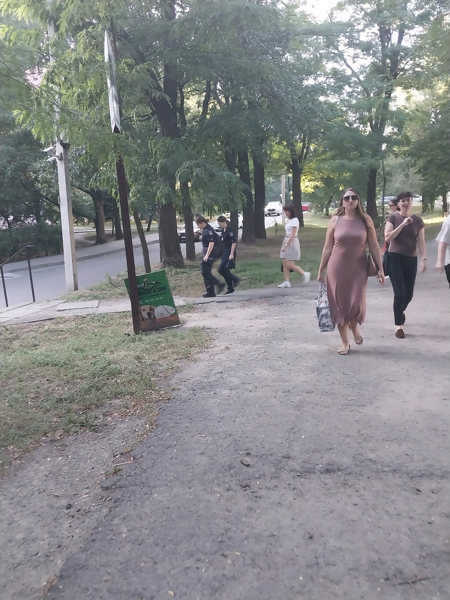 On a cloudy day in Dnipro, Ukraine, two police officers in dark uniforms walk to the right on a paved path. Behind them, a woman in a light-colored dress walks to the right, carrying a shopping bag and wearing sunglasses. Further behind and to the right, another woman in a dark top and black pants walks to the right, and a third person in a light-colored shirt is partially visible walking to the right. In the background, trees and a street with parked cars are visible. A green sign with text and images of dogs is placed on the ground to the left of the path.