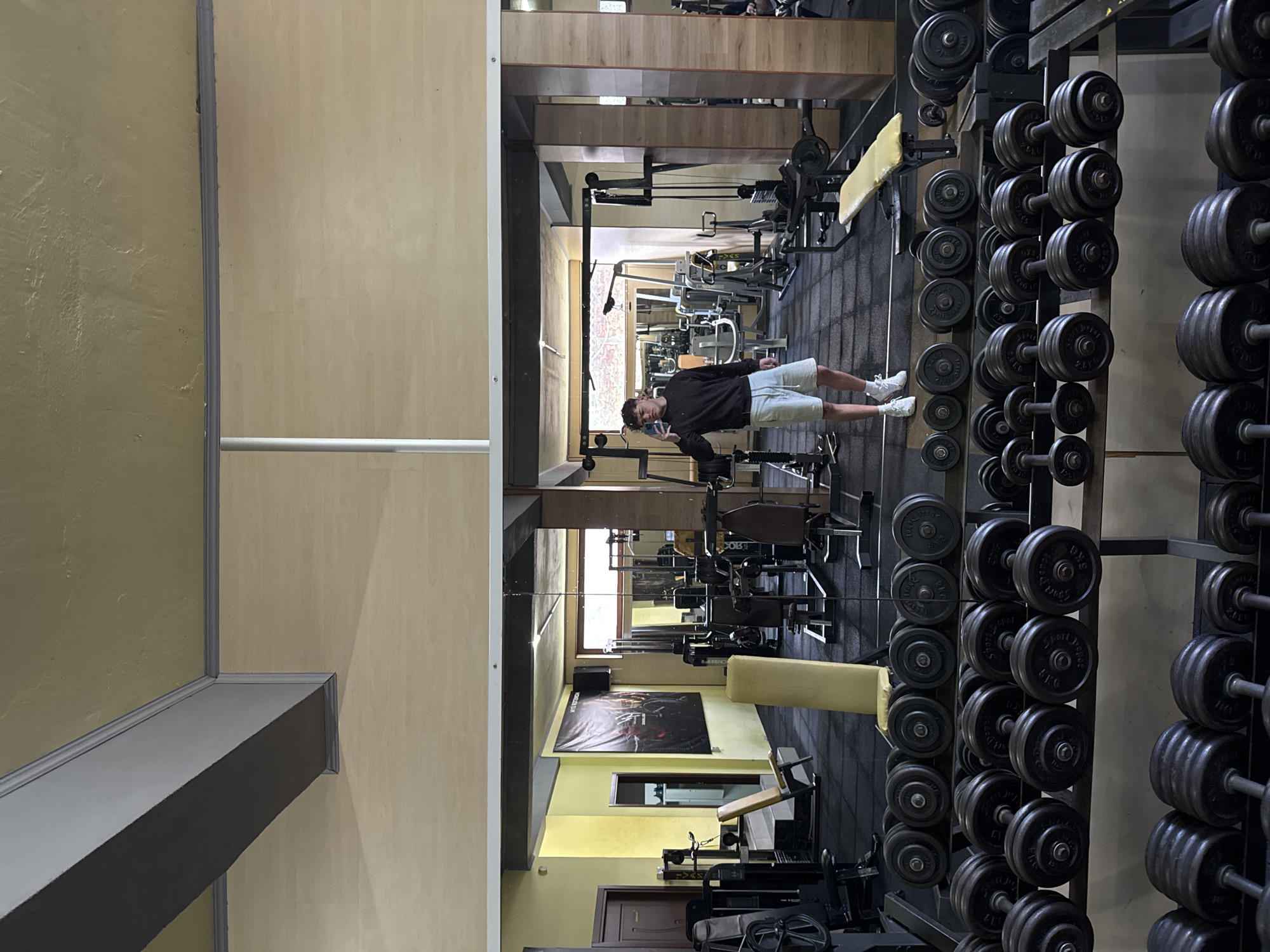 The photograph captures a gym interior. A young man is positioned in the central area, holding a phone, presumably taking a photograph. The gym contains rows of dumbbells, arranged on metal racks to the right of the frame. Various gym machines are visible in the central area, including weight benches, cable machines, and other fitness equipment. The gym has a modern aesthetic, with a black rubberized floor and neutral-colored walls. A poster is affixed to the wall. The location is a gym in the city of Goce Delcev, Bulgaria.