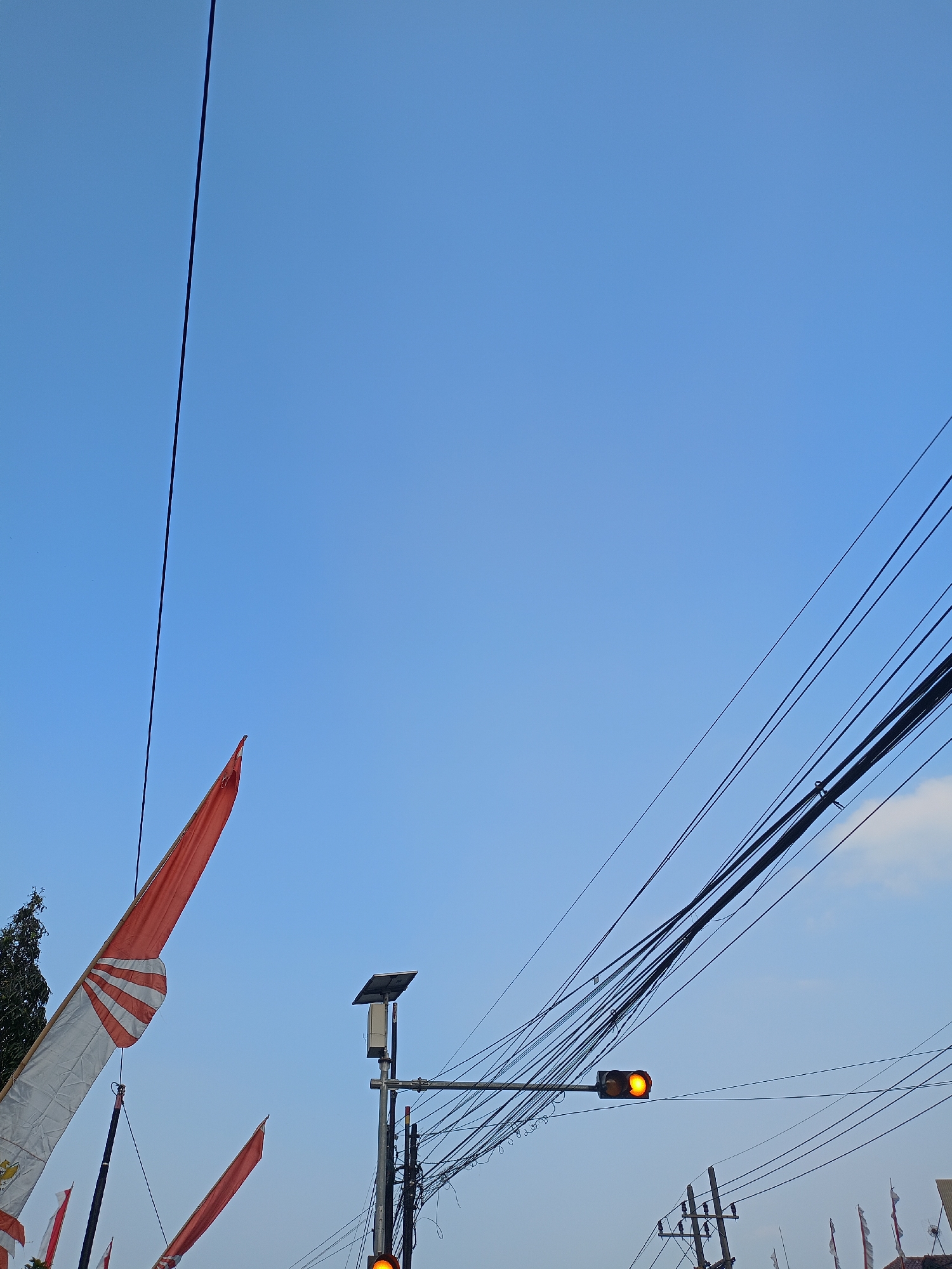The image captures a clear blue sky in Batu, Indonesia, with a collection of overhead wires dominating the right side of the frame. A traffic light shows a lit yellow signal. In the foreground, two red and white flags are visible, one larger and partially unfurled on the left, and another smaller one further to the right. A street light with a solar panel is also present. The scene suggests an urban or semi-urban environment.