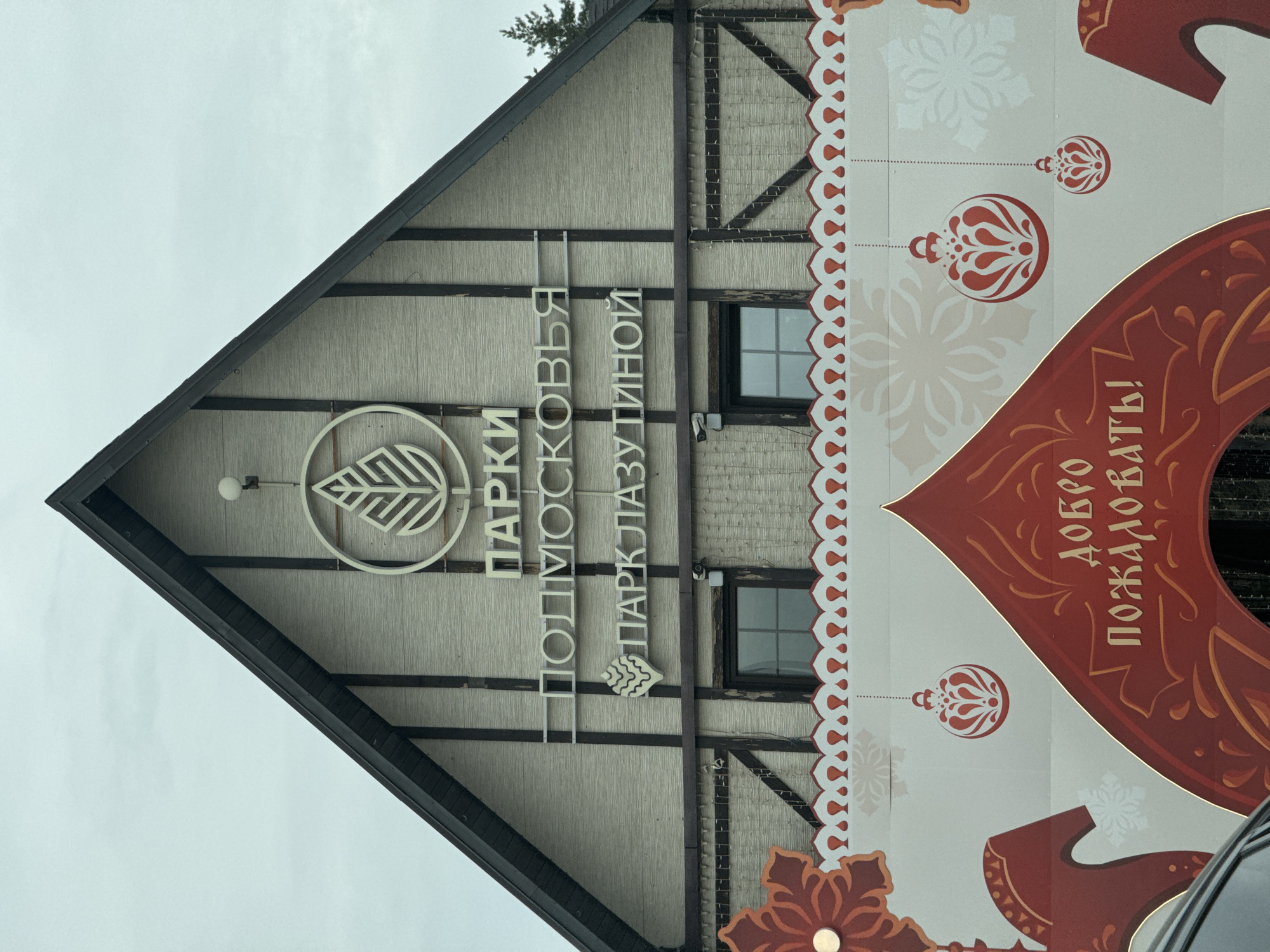The image depicts the exterior of a building at Lazutina Park (Парк Лазутиной) in Odintsovo, Moscow Oblast, Russia, under an overcast sky. The building features an A-frame gabled facade constructed with light beige textured paneling and dark brown decorative beams. Prominent white signage on the gable displays a stylized leaf logo, the text "ПАРКИ ПОДМОСКОВЬЯ" (Parks of Moscow Oblast), a stylized pinecone logo, and "ПАРК ЛАЗУТИНОЙ" (Lazutina Park). A small white spherical light fixture is mounted on the upper left side of the gable. Two dark-framed, multi-pane windows are visible below the main signage, with a small dark security camera mounted above the left window frame.
In the foreground, a large, white decorative banner or gate partially obscures the lower section of the building. This banner features a central red and gold arched structure with the Cyrillic text "ДОБРО ПОЖАЛОВАТЬ!" (Welcome!). The top edge of the banner has a red and white scalloped pattern. The white background of the banner is adorned with light gray snowflake patterns and several red and white stylized ornamental baubles hanging from dotted lines. Red and orange decorative elements, suggestive of traditional Russian folk art, are visible on the left and right sides of the banner. A dark vehicle is partially visible in the bottom-left corner of the frame. A dark evergreen tree is partially visible behind the building's right roofline. No individuals are present in the scene.