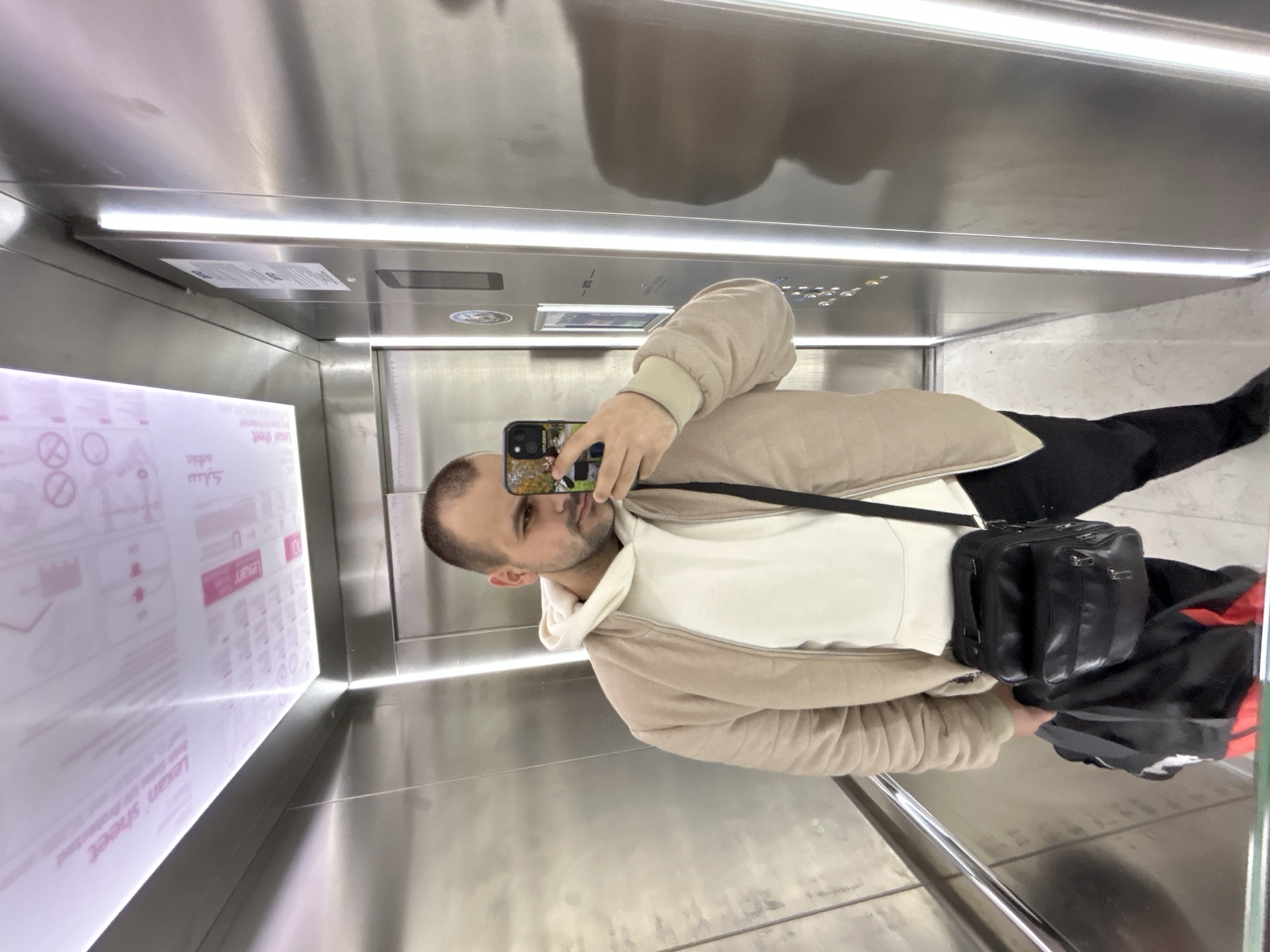 The image is a selfie taken by a light-skinned man with short, dark hair, wearing a light beige jacket, white hoodie, and dark pants. He is standing inside a metallic elevator. He is holding a mobile phone with a patterned case to take the picture. He has a black bag slung over his shoulder. The elevator interior is stainless steel with horizontal lighting fixtures. There is a sign on the left wall of the elevator that reads "Lexan sheet." The buttons and display panel of the elevator are visible above the man's head. The reflection of the elevator's ceiling is visible above the elevator buttons. There is no indication of the city of Plovdiv, Bulgaria in the image.