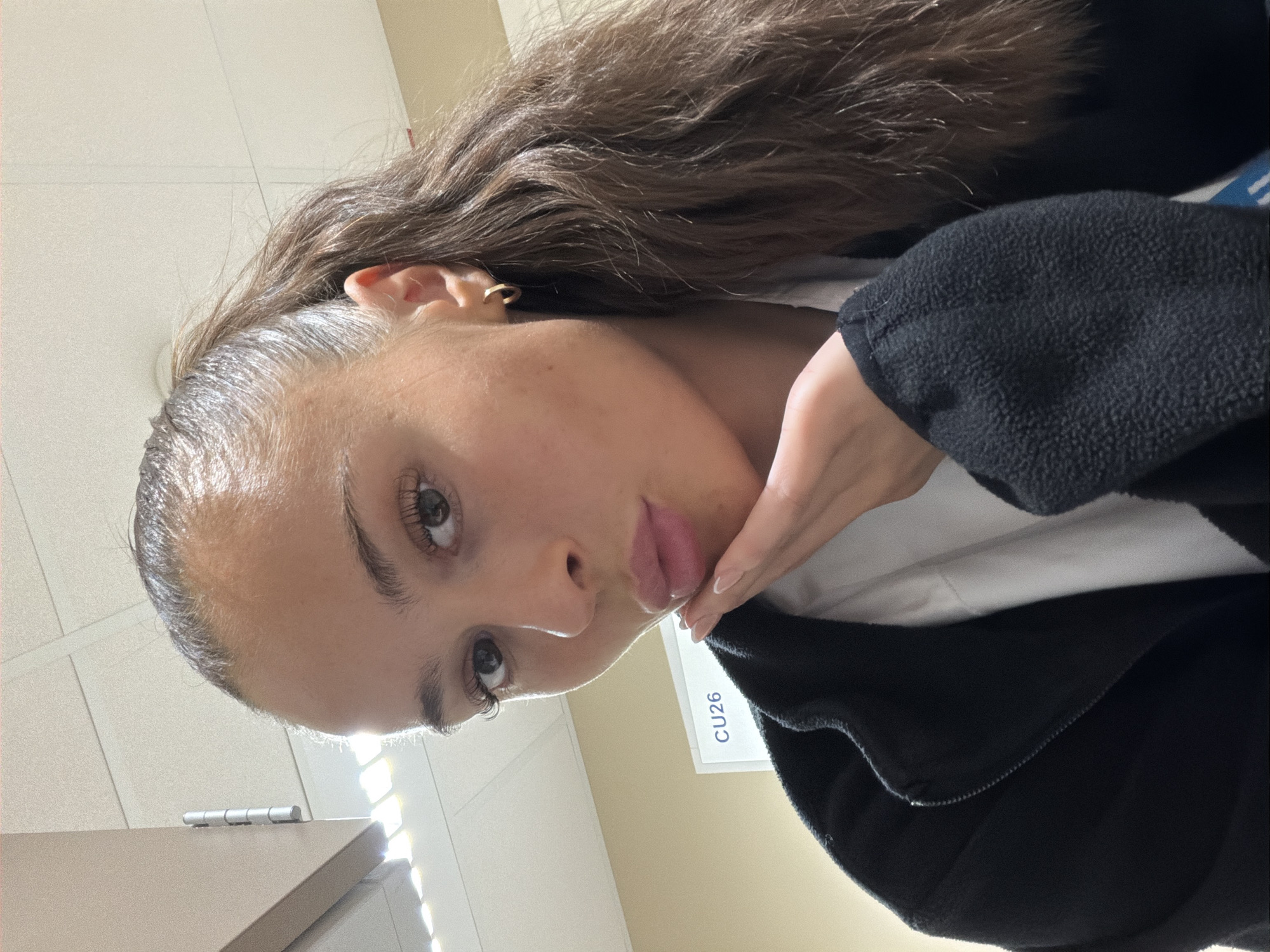 A young woman with long, dark brown hair pulled into a high ponytail, is shown in a close-up selfie. She is wearing a black zip-up jacket over a white t-shirt and has a gold hoop earring in her visible ear. Her right hand is resting on her chin, and her lips are pursed. In the background, a sign with "CU26" is visible on a beige wall, and ceiling tiles with fluorescent lighting are overhead. There are no visible indications of the location being in Harstad, Norway.