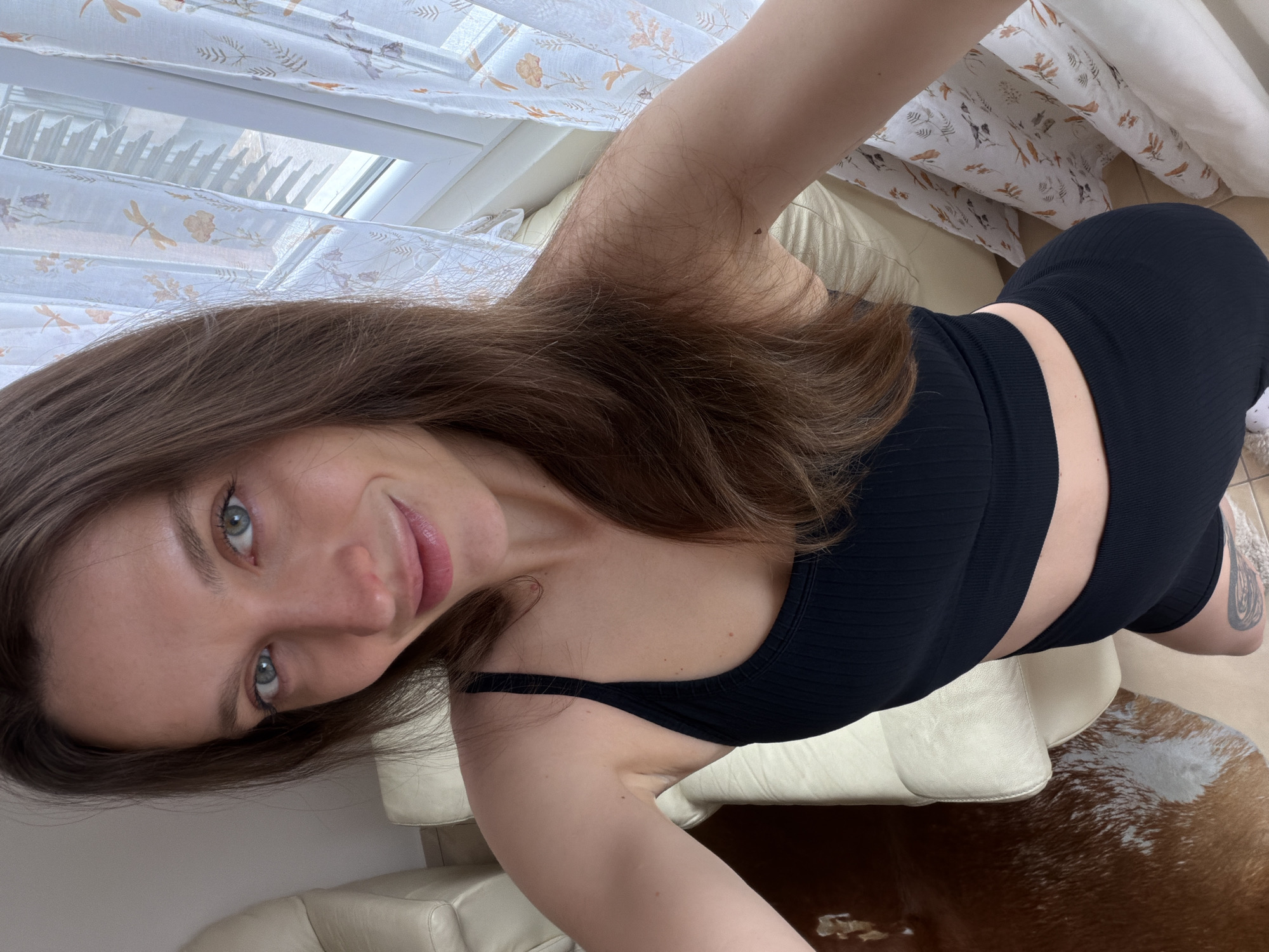 A woman is posing for a selfie indoors. She has long brown hair, fair skin, and blue eyes. She is wearing a black crop top and shorts. She is sitting on a light-colored leather sofa. Behind her is a window with a patterned curtain. The floor is covered with a brown and white cowhide rug. No information to determine if the photo was taken in Budva, Montenegro, or elsewhere is visible in the image.