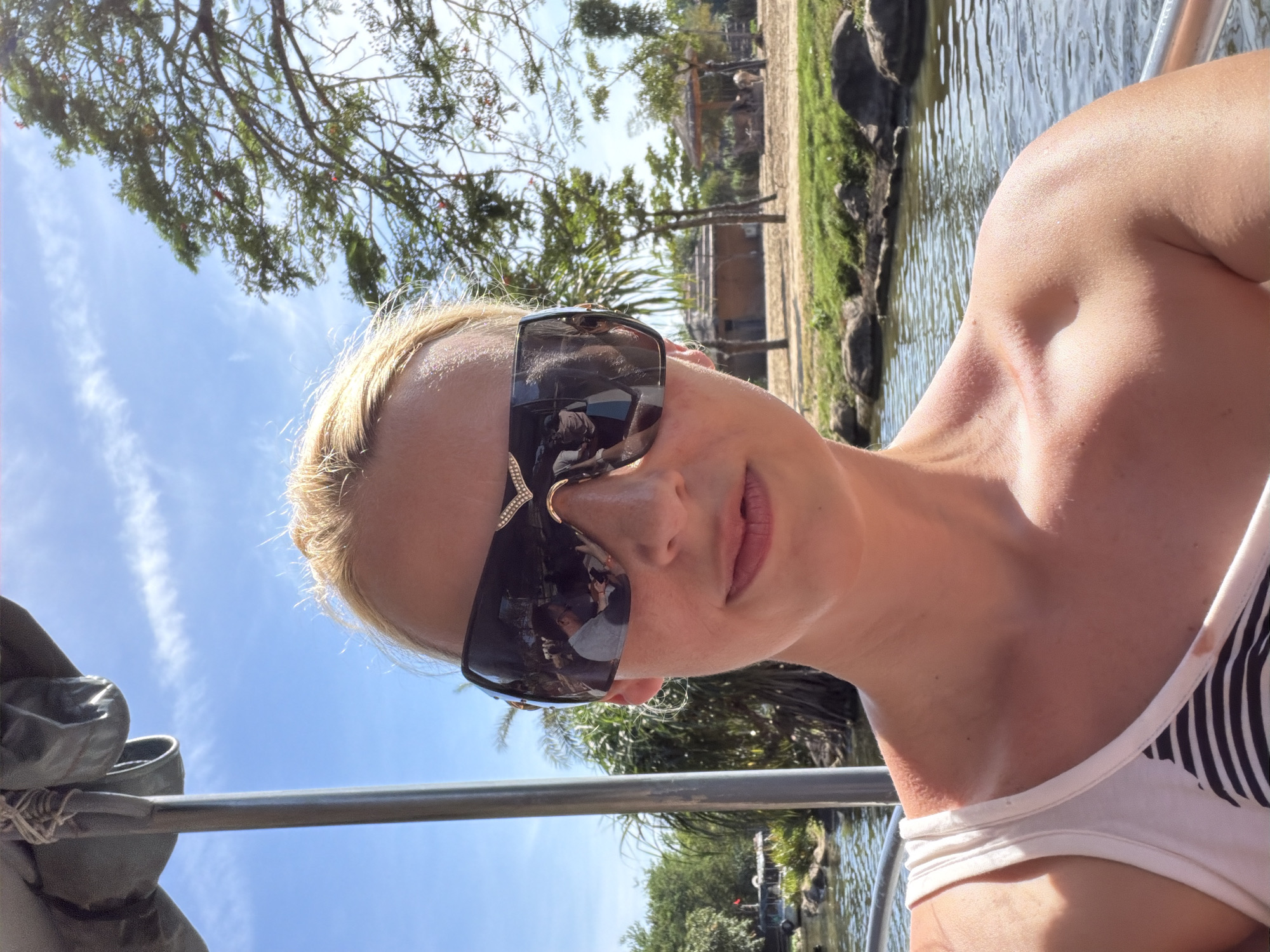 A woman with fair skin and blonde hair, wearing sunglasses and a white tank top with black stripes, is the primary subject of the image. She appears to be on a boat. The background features a body of water, trees, and green vegetation. The sky is blue with some clouds. The reflection in the sunglasses shows other people on the boat. The environment suggests a tropical or subtropical location. There is not enough evidence to place the location in Đà Nẵng, Vietnam.
