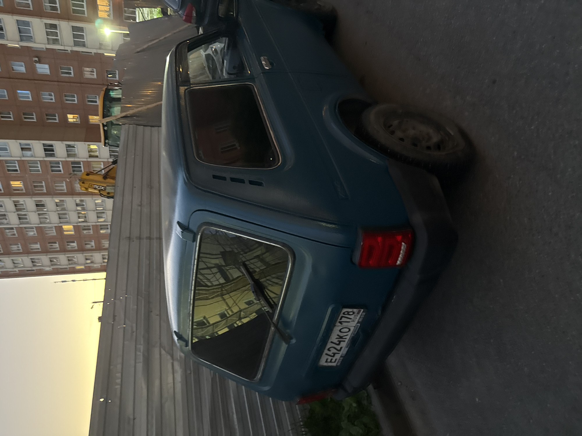 A teal-colored Lada Niva is parked on an asphalt surface, likely a street or parking lot, in front of a metal construction fence. Beyond the fence, a multi-story brick apartment building stands, its windows illuminated by what appears to be dusk or artificial lighting. A yellow backhoe loader is partially visible behind the fence, suggesting ongoing construction. The license plate on the Lada Niva reads "E424KO 178", indicating it is registered in the Saint Petersburg region of Russia. The sky above is a soft yellow and orange, characteristic of a sunset or sunrise. The overall scene depicts a common urban environment with elements of residential life and construction activity.