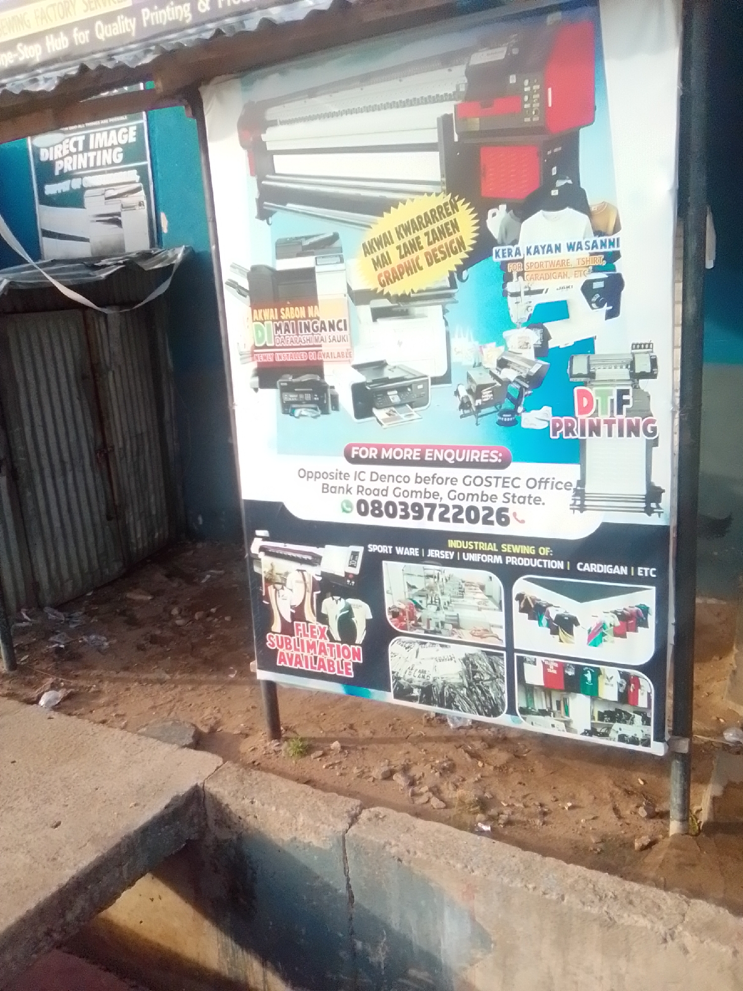 The image shows a street scene in Pantame, Nigeria, featuring a large advertisement sign for a printing and sewing business. The sign is colorful and displays various printing services such as "DIRECT IMAGE PRINTING," "DTF PRINTING," and "FLEX SUBLIMATION AVAILABLE." It also highlights "INDUSTRIAL SEWING OF: SPORT WARE | JERSEY | UNIFORM PRODUCTION | CARDIGAN | ETC." The advertisement includes images of printing machines, sewing machines, and various apparel items like t-shirts and jerseys.
The sign is propped up on metal poles and is positioned near a corrugated metal structure, possibly a shop or stall. The ground in front of the sign is dirt and rubble, with some debris scattered around. A concrete gutter or drainage system is visible in the foreground. The overall environment appears to be an outdoor commercial area, common in Nigerian towns. There are no people clearly visible in the image, but the advertisement implies a business operating in this location. The lighting suggests it is daytime, though the overall clarity is slightly reduced due to image quality.