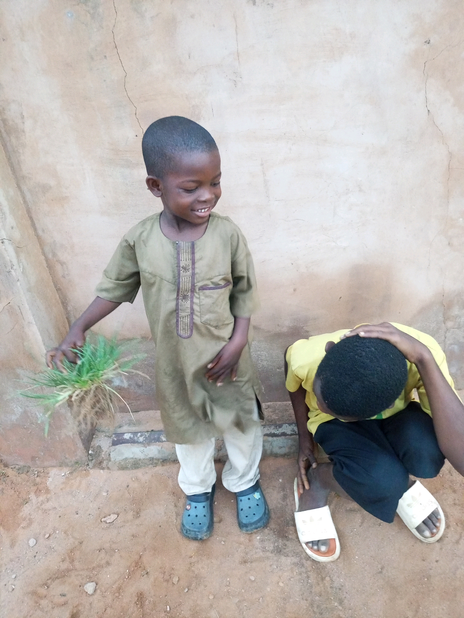 Two young boys are in an outdoor environment in front of a rough, beige-colored wall. The boy on the left is standing and smiling, holding a clump of grass in his left hand. He is wearing a light brown, long-sleeved tunic with purple stitching and light-colored pants, along with blue Croc-style shoes. The boy on the right is crouching and has his head bowed, with his right hand resting on his head. He is wearing a yellow t-shirt and dark pants, with white slides on his feet. The ground is sandy and dusty, with some small stones and debris. The wall behind them appears cracked and weathered. The scene is consistent with an outdoor setting in Tempure, Nigeria.