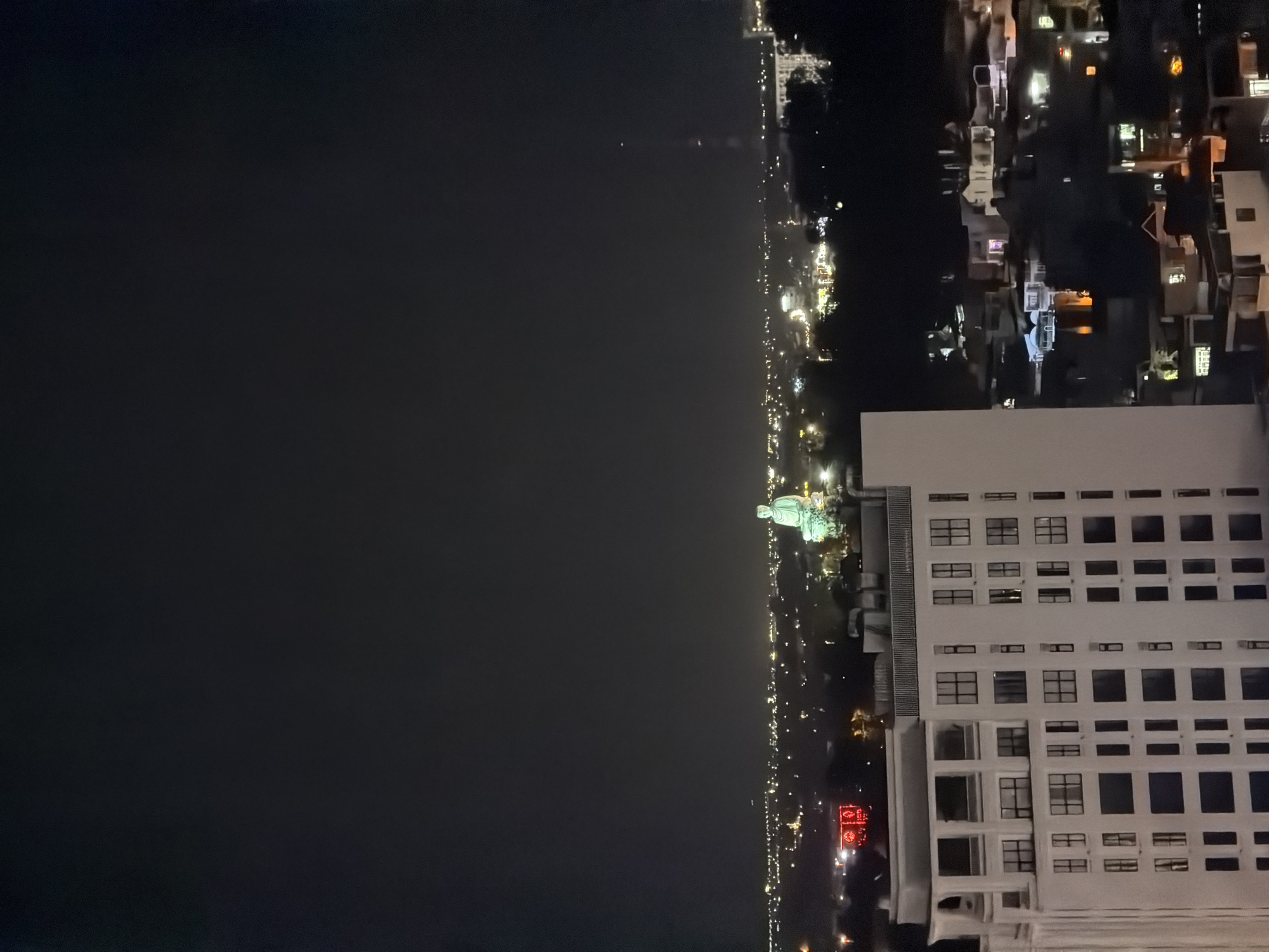 At night, the Ward of Nha Trang, Vietnam reveals its urban character, as seen from an elevated perspective. The dark expanse of the sea contrasts with the city's lights lining the coastline. A prominent white building with many windows stands out, positioned near other lit structures and houses. Notably, a statue stands aglow amidst the city's lights. The overall environment is urban, accentuated by the artificial illumination that defines the night scene.