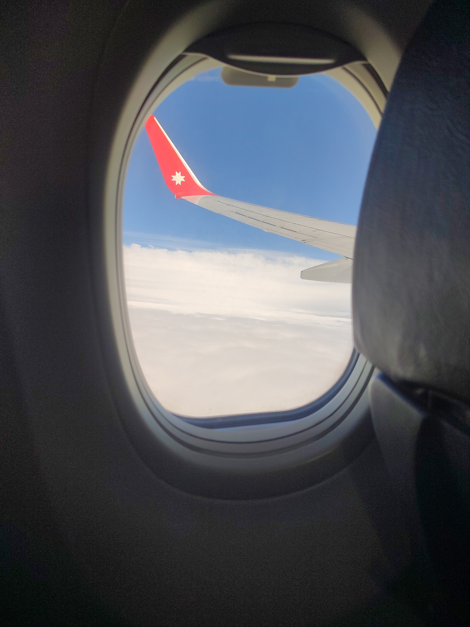 The image is a view from an airplane window. The aircraft's wing is visible, with a red wingtip that features a white star. Outside the window, there is a clear blue sky above a thick layer of white clouds. Inside the plane, part of a seat is visible next to the window. The location cannot be determined from the image, and therefore, it cannot be confirmed that this image depicts Chaikovsky, Russia.