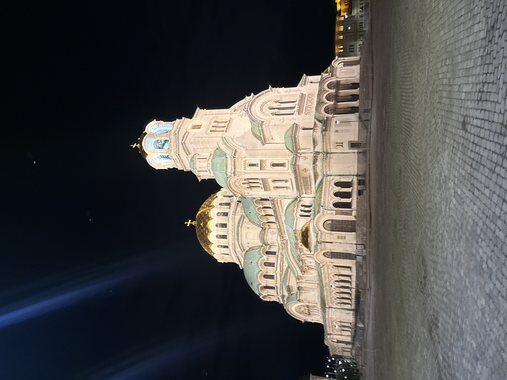 The image depicts the Alexander Nevsky Cathedral in Sofia, Bulgaria, at night. The cathedral is a large, ornate Orthodox cathedral with multiple domes, the largest of which is gilded. The building is illuminated by artificial lights, creating a dramatic contrast against the dark night sky. In the foreground, a cobblestone plaza is visible, with some streetlights and fences. The sky is dark and clear, with some faint light streaks that could be from artificial lights or possibly a long exposure effect. There are no people visible in the image. The architectural style is Neo-Byzantine, characteristic of many Russian Orthodox churches. The overall impression is one of grandeur and solemnity.