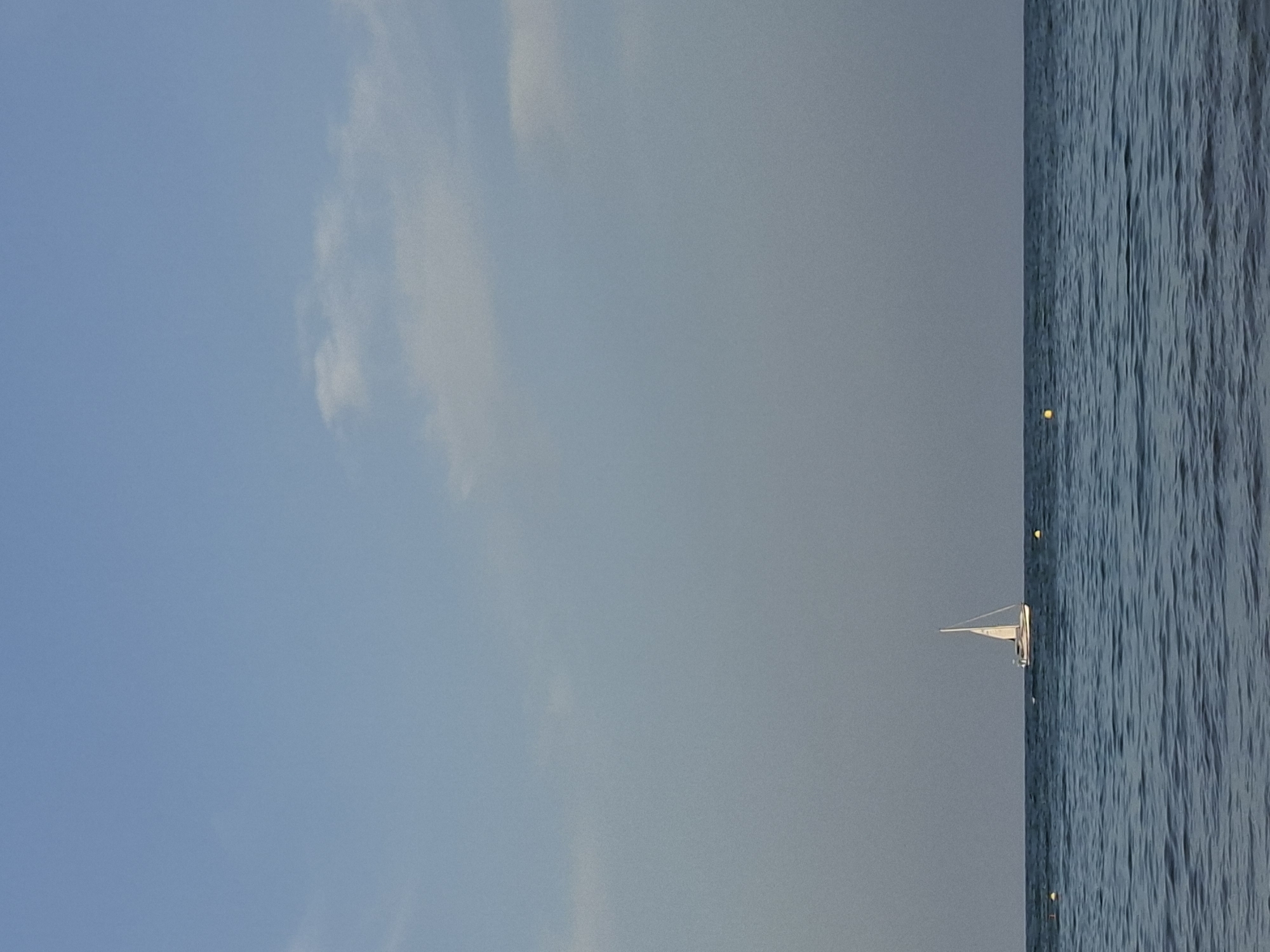 The image captures a serene seascape at the coast of Torrevieja, Spain. The horizon is clearly defined, separating the deep blue, gently rippling water from a pale blue sky with scattered, wispy clouds. A lone white sailboat with its sails unfurled is positioned in the middle ground, sailing on the calm sea. Three small yellow buoys are visible on the water's surface, marking a boundary or area. The scene is devoid of people and any activity, emphasizing a peaceful and expansive natural setting. The overall impression is one of tranquility and open space.
