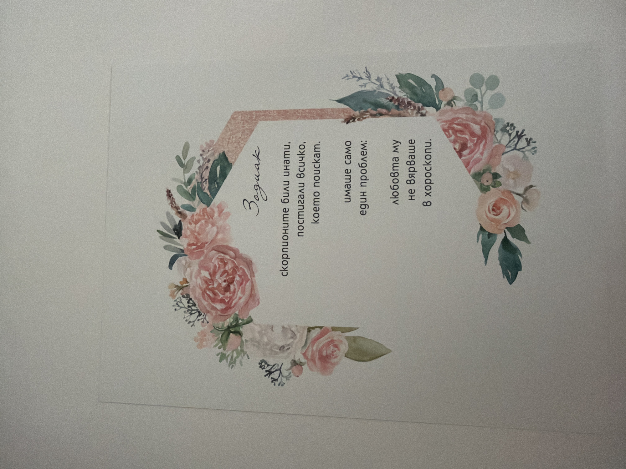 A card with a floral design and text in Bulgarian. The text reads "Зодиак" (Zodiac), followed by a quote. The quote talks about scorpios being smart and achieving everything they want, but having only one problem: their love did not believe in horoscopes. The card is on a plain, light-colored surface, possibly a wall or a table. There are no people or any other objects visible in the frame. There is no information to determine its location in Pazardzhik, Bulgaria.