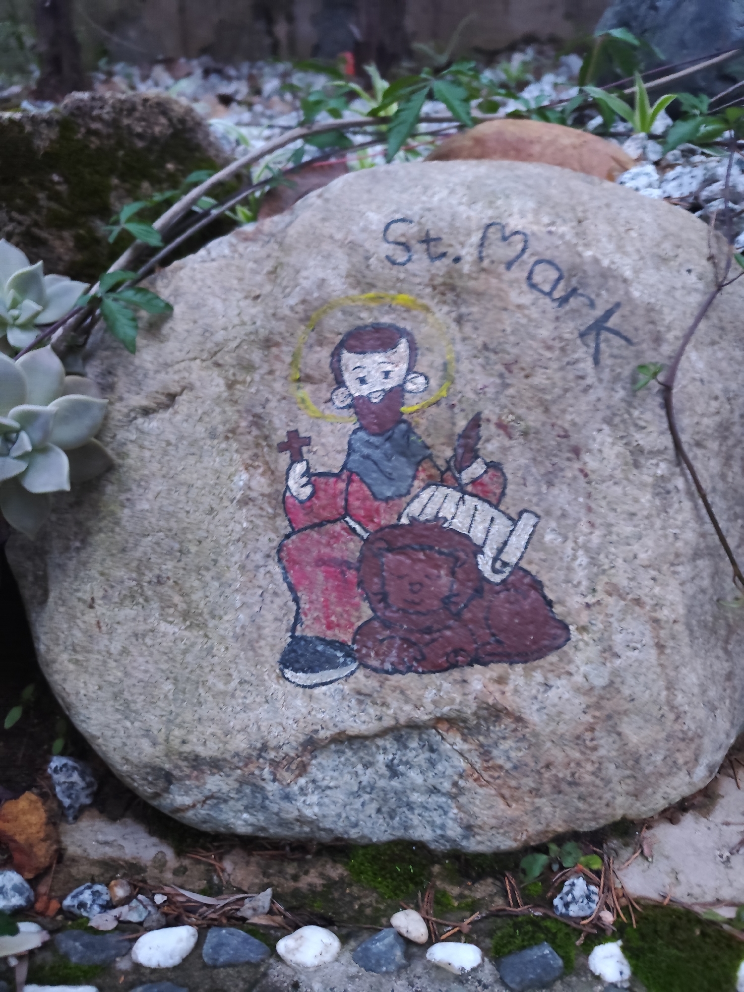 A medium-sized stone is the focal point of the image, featuring a painted depiction of St. Mark with a halo, holding a cross, and accompanied by a lion. The name "St. Mark" is written in black letters above the painted figure. The stone is situated in what appears to be a garden setting, surrounded by moss, small rocks, a succulent plant, and various green plants with vines. The scene is dimly lit, suggesting it might be outdoors in a shaded area. There is no explicit information in the image confirming that the location is in Da Lat, Vietnam.