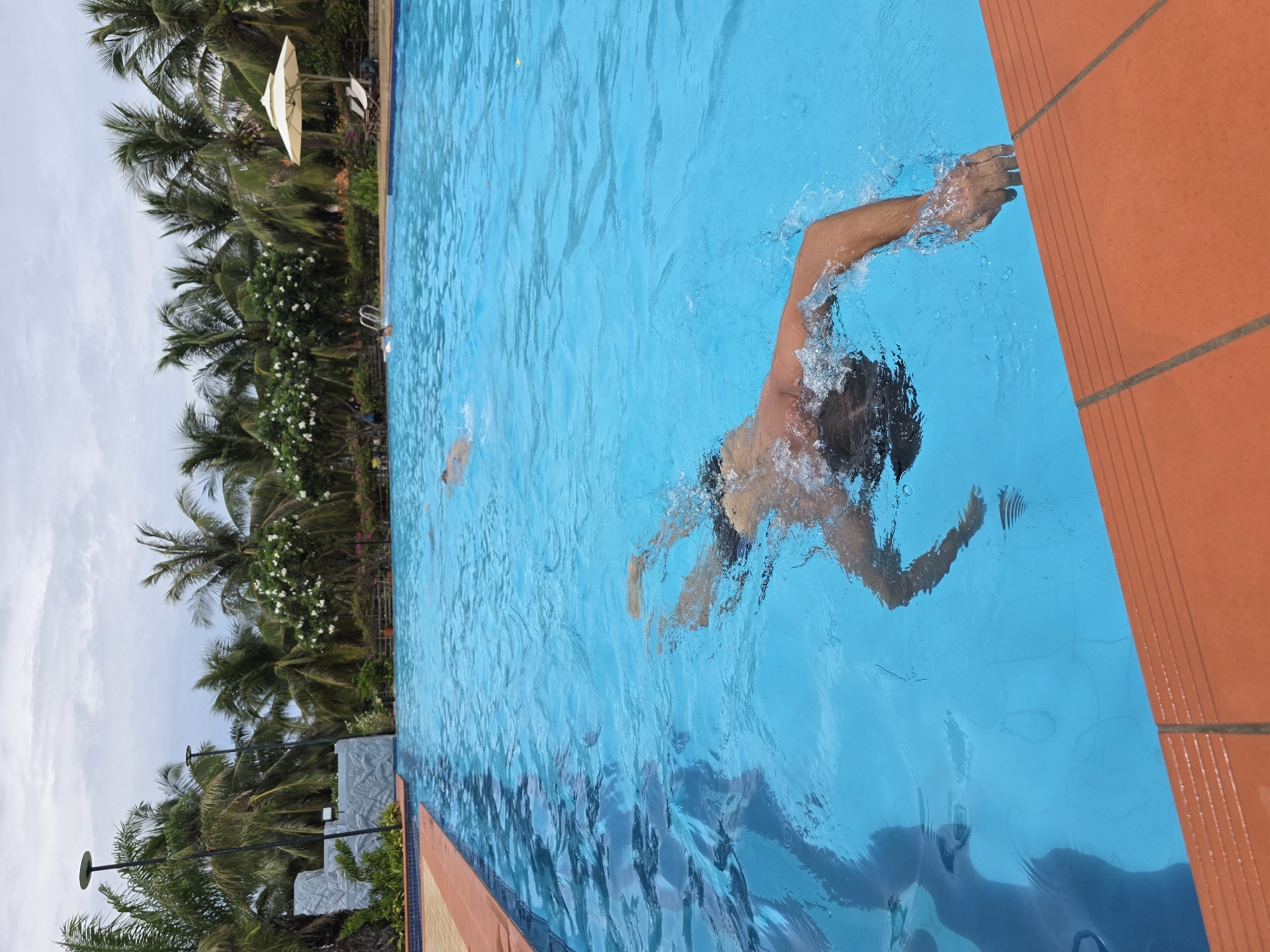 The image presents an overhead view of an outdoor swimming pool area, featuring a person actively swimming in bright blue water. The swimmer is captured mid-stroke, with their back partially visible above the water and one arm extended forward, creating splashes. Their hair appears dark. Another individual is faintly discernible in the distance within the pool.
The swimming pool is bordered by an orange tiled edge. Beyond the pool, a vibrant tropical landscape dominates the background. Numerous tall palm trees with lush green fronds line the area, interspersed with bushes bearing white flowers. A white outdoor parasol and a few white chairs are visible amongst the greenery, suggesting a leisure or lounging zone. Further down the poolside, outdoor lampposts are present. The sky above is overcast, indicating a cloudy daytime setting. The scene evokes a sense of relaxation and recreation, typical of a resort or hotel pool in a warm climate. No visible text is present in the image. The location is consistent with Phu Thuy Ward, Vietnam.