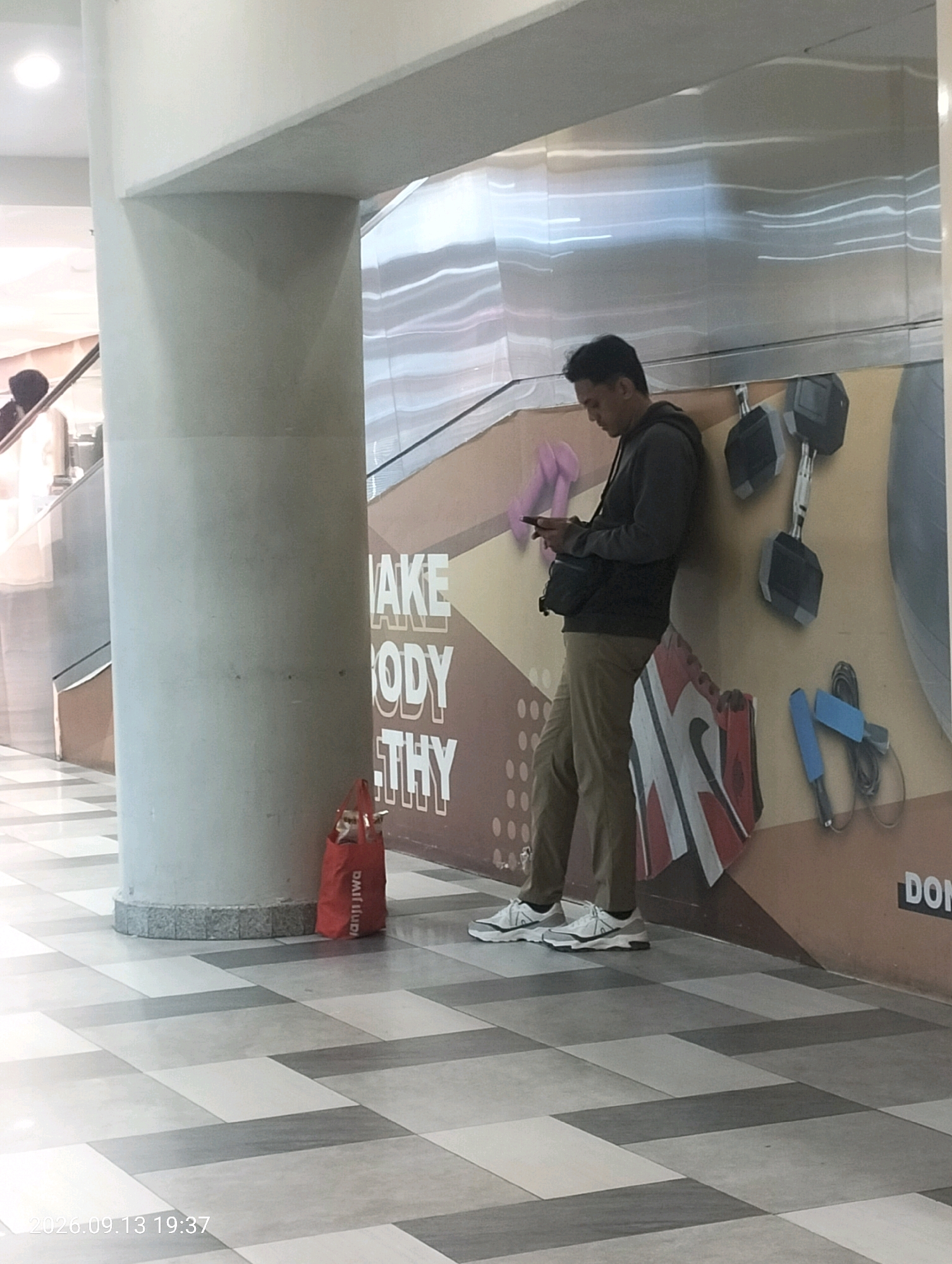 A young man stands leaning against a wall in a brightly lit, indoor public space, possibly a mall or a gym in Surabaya, Indonesia. The wall behind him features a large, graphic design with text that appears to say "MAKE BODY HEALTHY," accompanied by images of gym equipment such as dumbbells and a jump rope. The man is dressed in a dark hooded sweatshirt, khaki pants, and white athletic shoes. He is looking down at his mobile phone, which he holds in his hands. A black crossbody bag is slung over his shoulder. To his left, a bright red shopping bag rests on the checkered tiled floor. The floor is made of large square tiles in alternating shades of grey and white, creating a geometric pattern. In the background, a staircase with reflective metal siding is visible, along with part of an escalator. The overall environment suggests a modern, public area designed for activity or leisure.