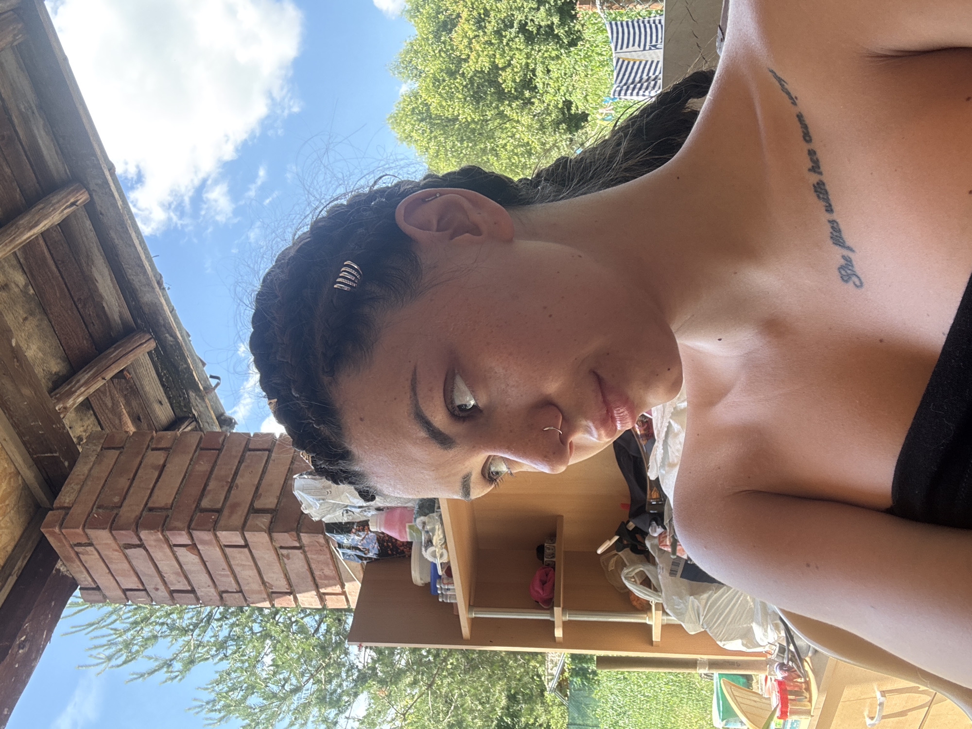 A young woman with braided hair and nose and ear piercings is pictured outdoors. She is looking to her left, with her shoulder and upper chest visible. A tattoo is visible on her upper chest, which reads "She flies with her own wings." Behind her, a wooden structure with a brick column is visible, along with shelves filled with various items. Trees and a blue sky with clouds are in the background. The scene appears to be a residential outdoor area, possibly a patio or garden. The image was taken in Belopoptsi, Bulgaria.