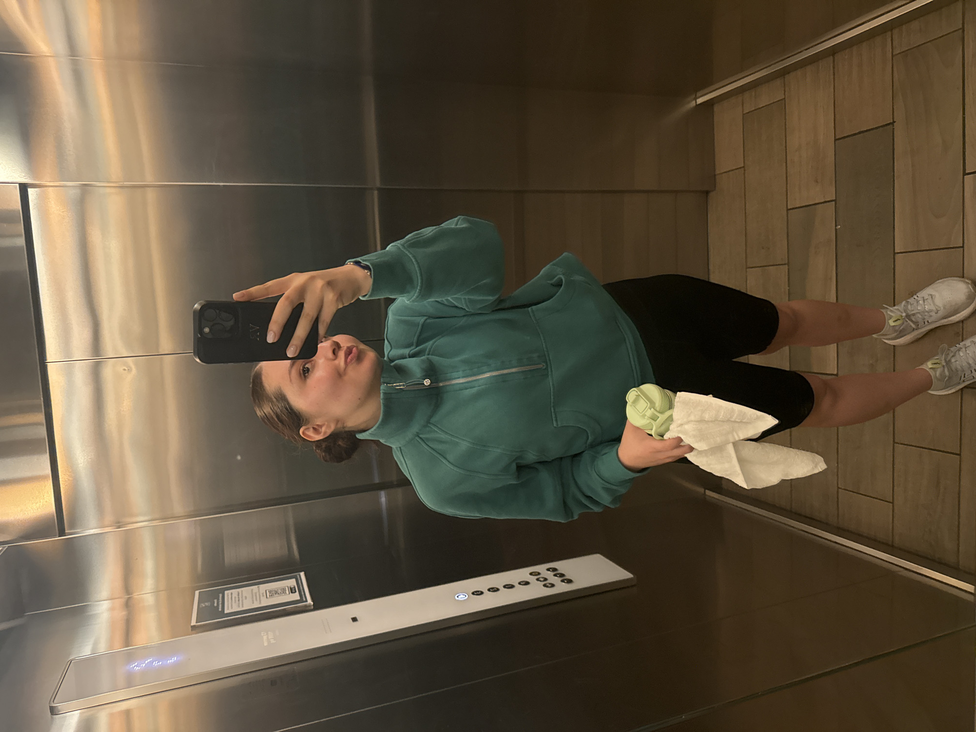 A woman is taking a selfie in an elevator. She is holding a phone in her left hand and a green bottle wrapped in a white towel in her right hand. The woman is wearing a green quarter-zip sweatshirt and black shorts. She has her hair pulled back in a bun and is wearing tennis shoes. The elevator has stainless steel walls and tan tiled floors. The elevator button panel is on the wall. There is a sign next to the elevator button panel. The location is unknown, however, the city is purported to be Mexico City, Mexico.