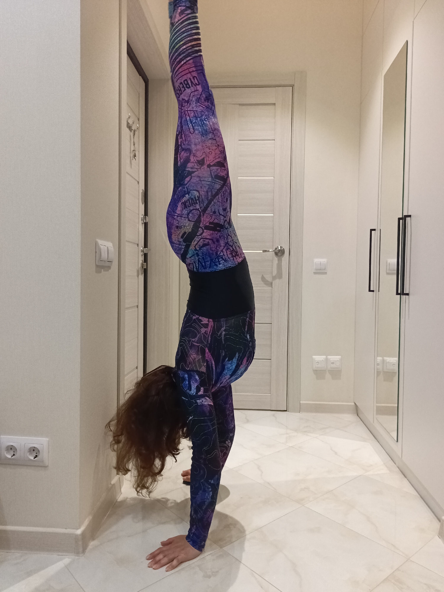 A woman with curly brown hair is performing a handstand indoors. She is wearing a full-body suit with a multi-colored, patterned design. Her body is in a straight line, supported by her hands which are placed on the floor. Her legs are extended upwards and her feet are not visible. The background consists of an interior hallway with light-colored walls and a tiled floor. There are several doors, electrical outlets, and a wardrobe with mirrored doors. The scene appears to be in a residential setting.