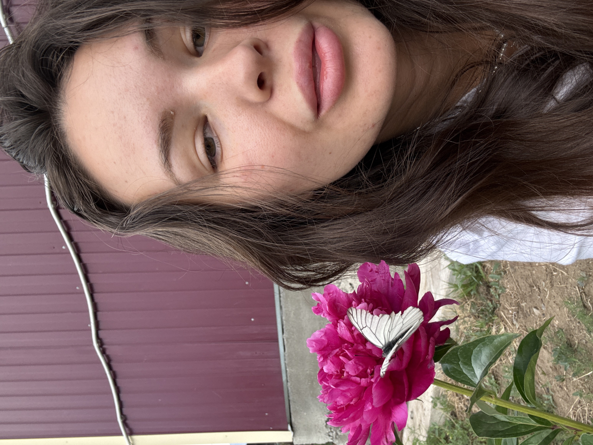 A young woman with long, dark brown hair takes a selfie outdoors, framing her face on the right side of the image. She is wearing a white top and a delicate silver necklace. Her skin shows some texture and minor blemishes. Her lips are full and slightly parted, with a hint of pink. Her eyes are partially closed, and she appears to be looking away from the camera. In the foreground, on the left side of the frame, a large, vibrant pink peony flower is in full bloom. A white butterfly with black markings rests on one of the peony's petals. Behind the woman and the flower is a burgundy corrugated metal fence, part of a building structure, with a white cable running horizontally across it. The ground below the flower is covered in dry soil and sparse green vegetation, consistent with an outdoor setting in Bely Yar, Russia.