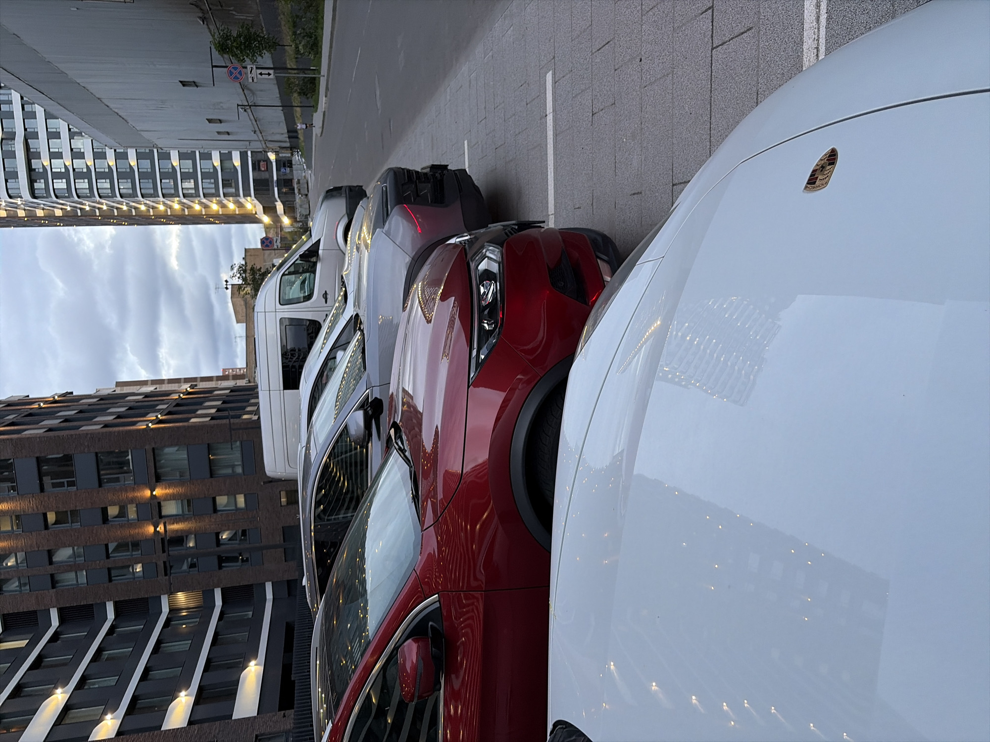 This is a medium-angle, eye-level shot of a line of parked cars on a city street, likely in Moscow, Russia. In the foreground, the hood of a white car, possibly a Porsche due to the visible emblem, is prominently featured. Behind it, a red SUV is parked. Further back, a grey SUV and a white van are visible, aligned along the street. The backdrop consists of modern, tall buildings with reflective windows, characteristic of urban architecture. The street is paved with cobblestones, and a sidewalk is present. The sky is overcast with clouds. There are no people clearly visible in the frame, only a distant silhouette of a person walking on the right side of the road. The lighting suggests it is either early morning or late evening.