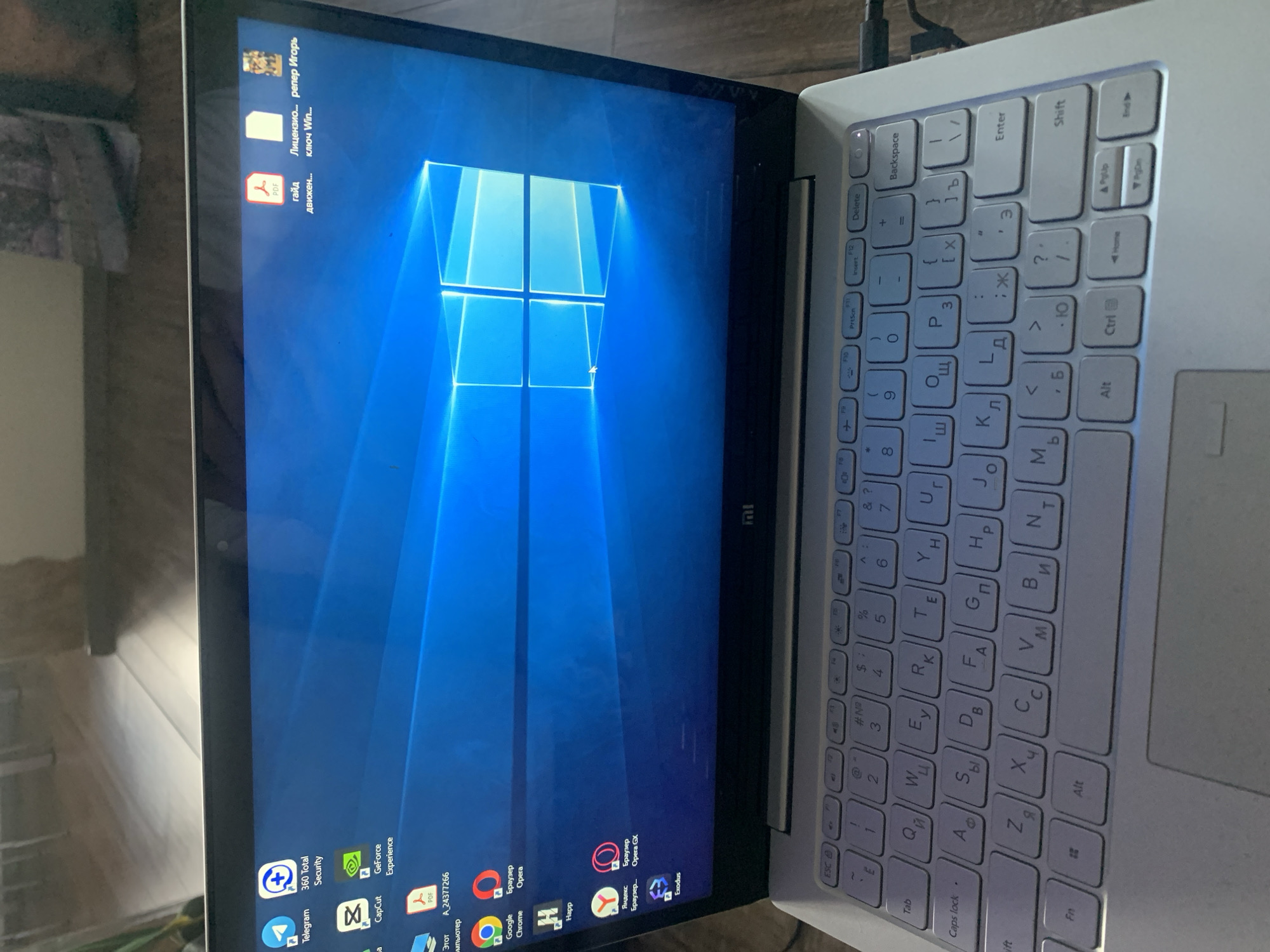 The image shows a laptop with a blue Windows wallpaper. Several icons are visible on the desktop, including Telegram, 360 Total Security, CapCut, GeForce Experience, a PDF file, and others. The keyboard is partially visible, and some of the keys have Cyrillic characters on them. A power cable is plugged into the side of the laptop. The background suggests an indoor environment, possibly a room. There is no indication of the city of Buzlanovo, Russia, in the image.