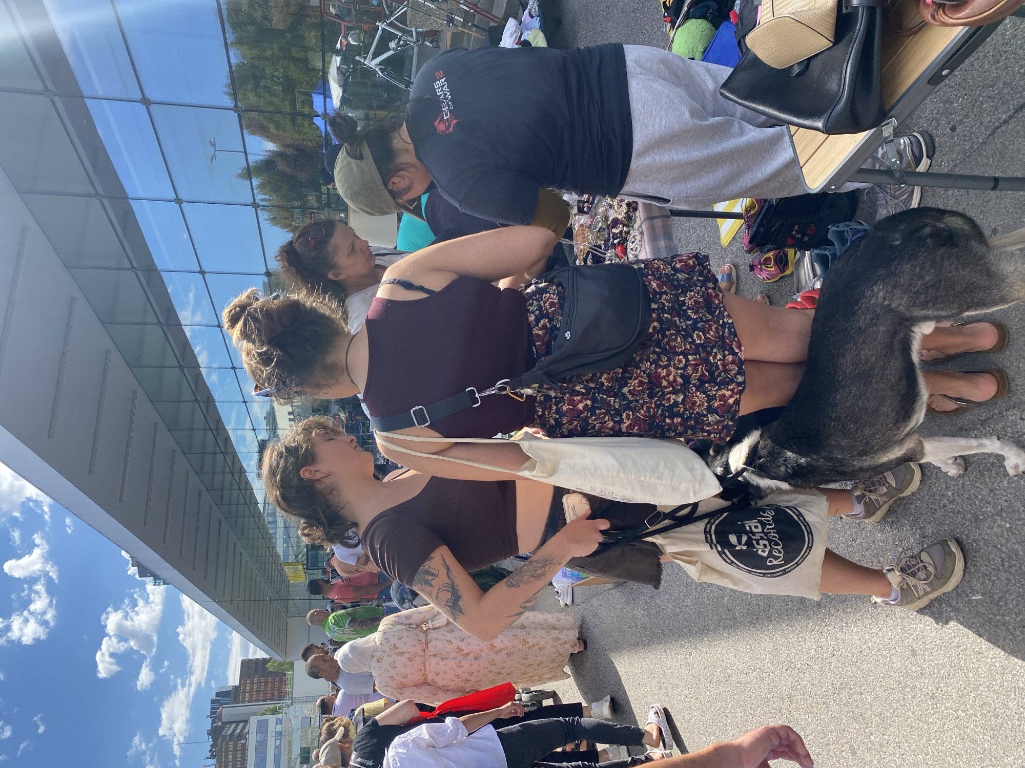 A group of people are gathered outdoors in front of a modern building with a glass facade. The scene appears to be a market or flea market, with tables displaying various items, including jewelry and clothing. In the foreground, a husky-type dog is standing between two women. One woman, wearing a brown crop top and a floral skirt, is holding a canvas bag with a "Dissol Records" logo. The other woman, with dark hair pulled back, is wearing a burgundy top and a patterned skirt. A third woman, wearing a green baseball cap and a black t-shirt with "Gears of War" on the back, is looking at items on a table. Other individuals are visible in the background, including people browsing and walking. The environment suggests a sunny day with a clear blue sky and some clouds. The architecture of the building indicates an urban setting, possibly in Vienna, Austria, given the context provided.