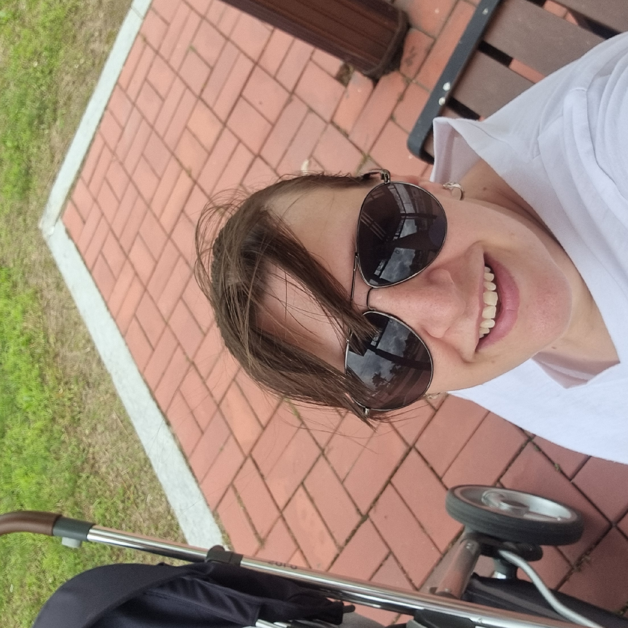 In an outdoor setting with red brick paving and green grass, a woman wearing aviator sunglasses and a white collared shirt smiles at the camera. Her brown hair is windswept. To her left, a portion of a dark-colored stroller with a silver frame and a brown handle is visible, along with its silver wheel. In the background, a dark brown wooden bench is partially visible on the right side. The image appears to be taken from a close-up, selfie-style perspective. The location is identified as Dunnae-myeon, South Korea.