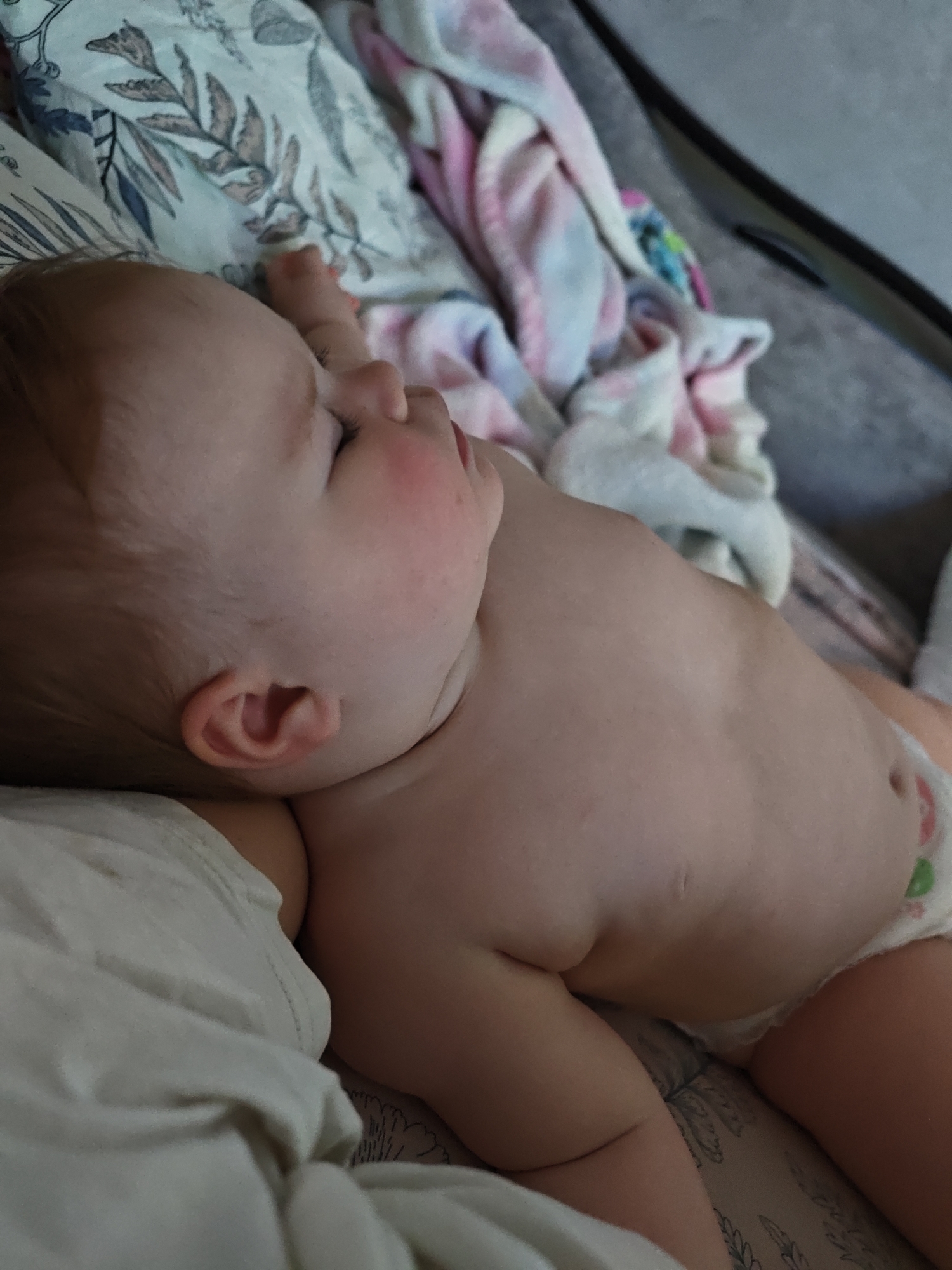 A baby is lying on its side on a patterned surface that appears to be a bed or a changing pad. The baby is wearing a diaper. The background includes a patterned pillow and a pink and white towel. The image was taken indoors. There is no evidence to confirm the location is in Chaikovsky, Russia.