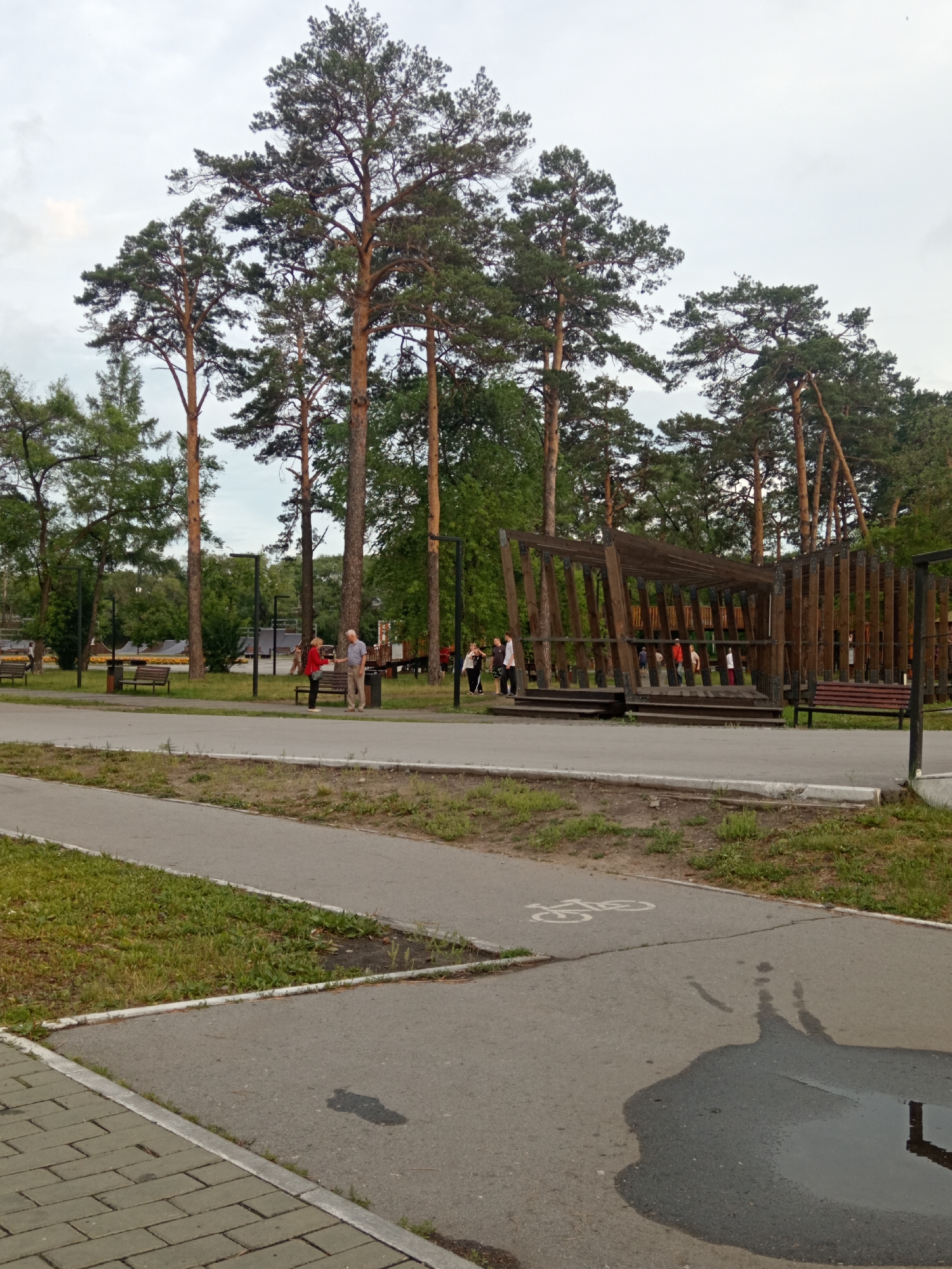 The image depicts an outdoor park scene. Several tall, mature pine trees are prominent in the background, with a cloudy sky overhead. In the midground, a group of people are visible near a wooden structure that appears to be an open-air pavilion or stage. To the left of this structure, two individuals, a man and a woman, are standing and conversing. Further left, several park benches are situated along a paved pathway. In the foreground, a paved path curves through the park, with a bicycle lane marking visible on the surface. A large puddle reflects the surrounding environment. The overall impression is of a public recreational area in a city park setting. The mention of Belogorsk, Russia, suggests this is a location within that city.