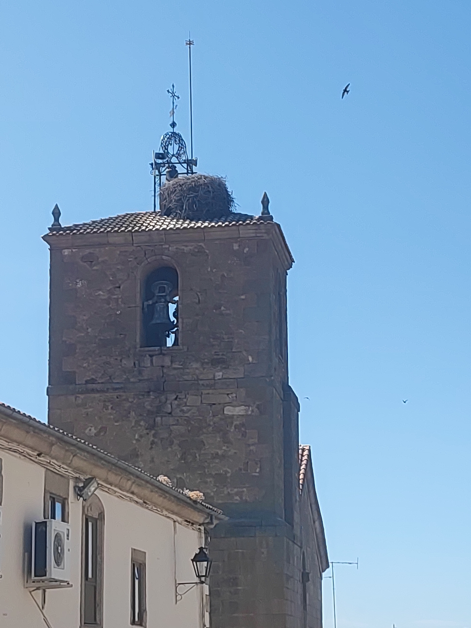 A clear, sunny day in Sobradillo, Spain, shows a close-up view of a stone church tower with a bell inside an arched opening. A large bird's nest is situated at the apex of the tower, just below a decorative metal structure topped with a cross. Several birds are visible in flight against the bright blue sky. In the foreground, the white facade of a building with a tiled roof is visible, featuring an air conditioning unit, windows, and a decorative lamppost. The image captures a slice of urban life and the natural environment.