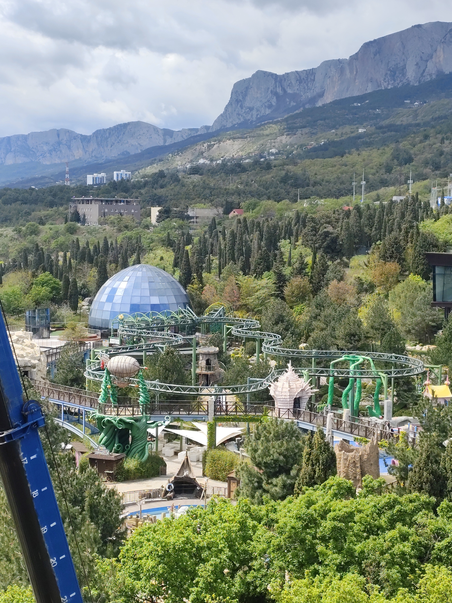 The image shows a park with a rollercoaster in the city of Unknown, Unknown. A blue-domed building is in the center of the park. The rollercoaster is green and winds through the trees and park features. There are trees and a building visible in the background. Mountains are present in the distance under a cloudy sky. Part of a blue crane is visible in the bottom left corner of the image.