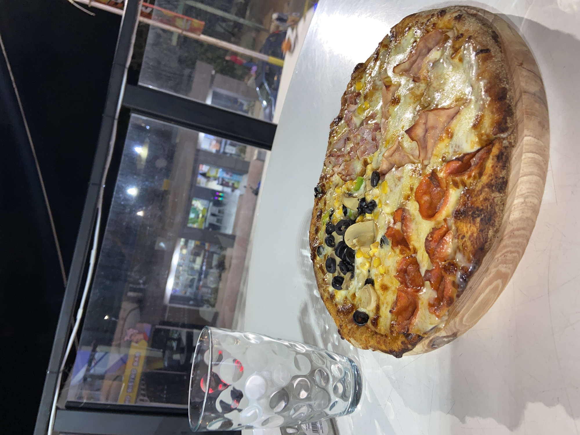 The image captures a large, appetizing pizza placed on a round wooden peel or cutting board, resting on a white table. The pizza is prominently featured in the right half of the frame, showcasing a thick, well-baked crust and distinct sections of different toppings.
The pizza appears to be divided into four quarters: one section features slices of ham or bacon with melted cheese; another section is topped with corn kernels, black olives, and sliced mushrooms; a third section is generously covered with pepperoni slices and cheese; and the fourth section also features ham or bacon with cheese, similar to the first, suggesting a popular combination of flavors.
In the foreground, to the left of the pizza, there is a clear glass with a distinctive textured surface, featuring an array of raised circular patterns. The white table surface shows some light scuffs or marks.
The background, visible through a large window, is dark, suggesting the photo was taken at night or in the evening. Reflections of interior lights and indistinct shapes of buildings or storefronts can be seen through the glass, indicating an urban or commercial setting. The general ambiance points to a restaurant or pizzeria.
The activity depicted is dining or about to dine, with the pizza ready for consumption. No people are visible, and no specific emotions are conveyed. While the setting is indoors, the specific location is not overtly indicated within the image itself, but the provided context places it in Juan Griego, Venezuela. Faint, illegible text is visible on a label beneath the glass and in some background reflections.