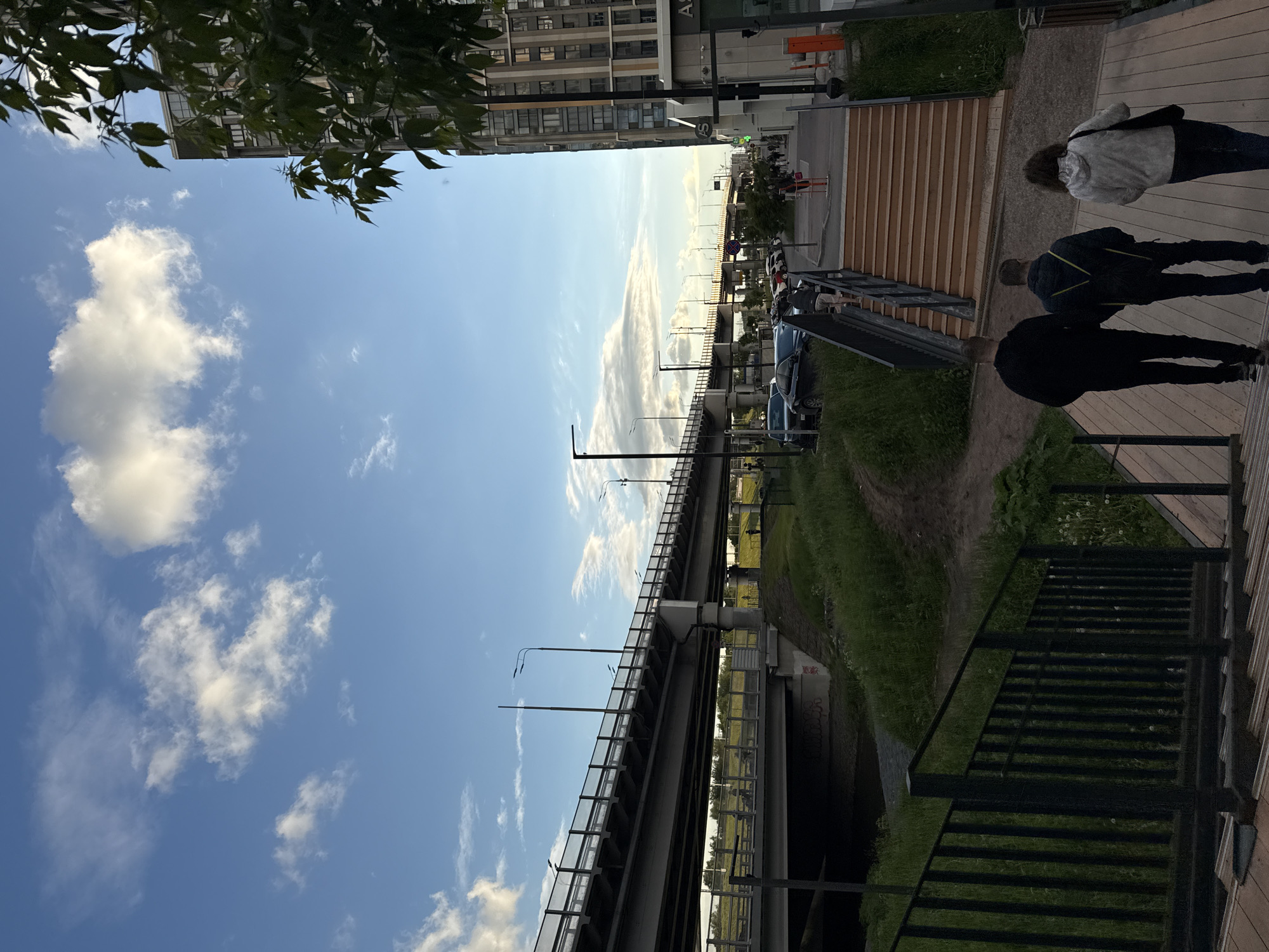 Captured from a low vantage point on a raised wooden platform, the image portrays an elevated view of a street scene in what appears to be Moscow, Russia. The photograph is rotated 90 degrees counter-clockwise.
Three individuals are walking along the wooden platform. Below the platform, a grassy area leads to an elevated roadway or overpass supported by pillars. Several parked cars are seen adjacent to the walkway. In the distance, buildings rise against a partly cloudy sky, interspersed with trees and foliage visible in the upper part of the image. Street lighting is evident along the elevated roadway.