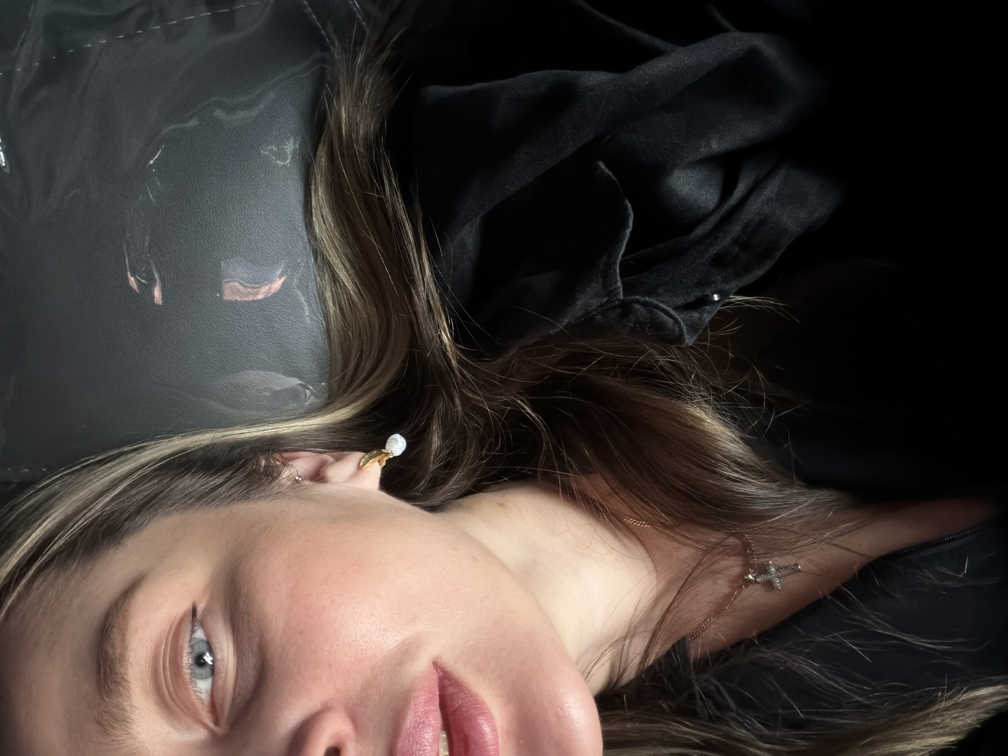 A close-up portrait shows a young woman lying down, her head turned towards the viewer, captured from a slightly overhead and rotated angle. Her light blue-grey eyes gaze directly, conveying a serene expression. Her lips are full and naturally colored in a soft pink hue. Long, wavy brown hair with distinct lighter streaks, possibly highlights, cascades around her face and extends into the dark background.
She wears a small golden hoop earring with a white, pearl-like bead on her left earlobe, and another tiny silver stud is visible higher on her ear. A delicate golden chain necklace with a small, sparkling cross pendant rests on her neck. She appears to be dressed in dark, possibly black, fabric, which is visible as crumpled folds around her head and upper body.
The background directly behind her head and to the left is a dark, smooth material, possibly leather or a similar synthetic fabric, showing subtle reflections and textures. The lighting is soft and diffused, creating an intimate and subdued mood. No specific activity is depicted other than posing for the portrait. There is no visible text in the image. The location context provided for this scene is Moscow, Russia.