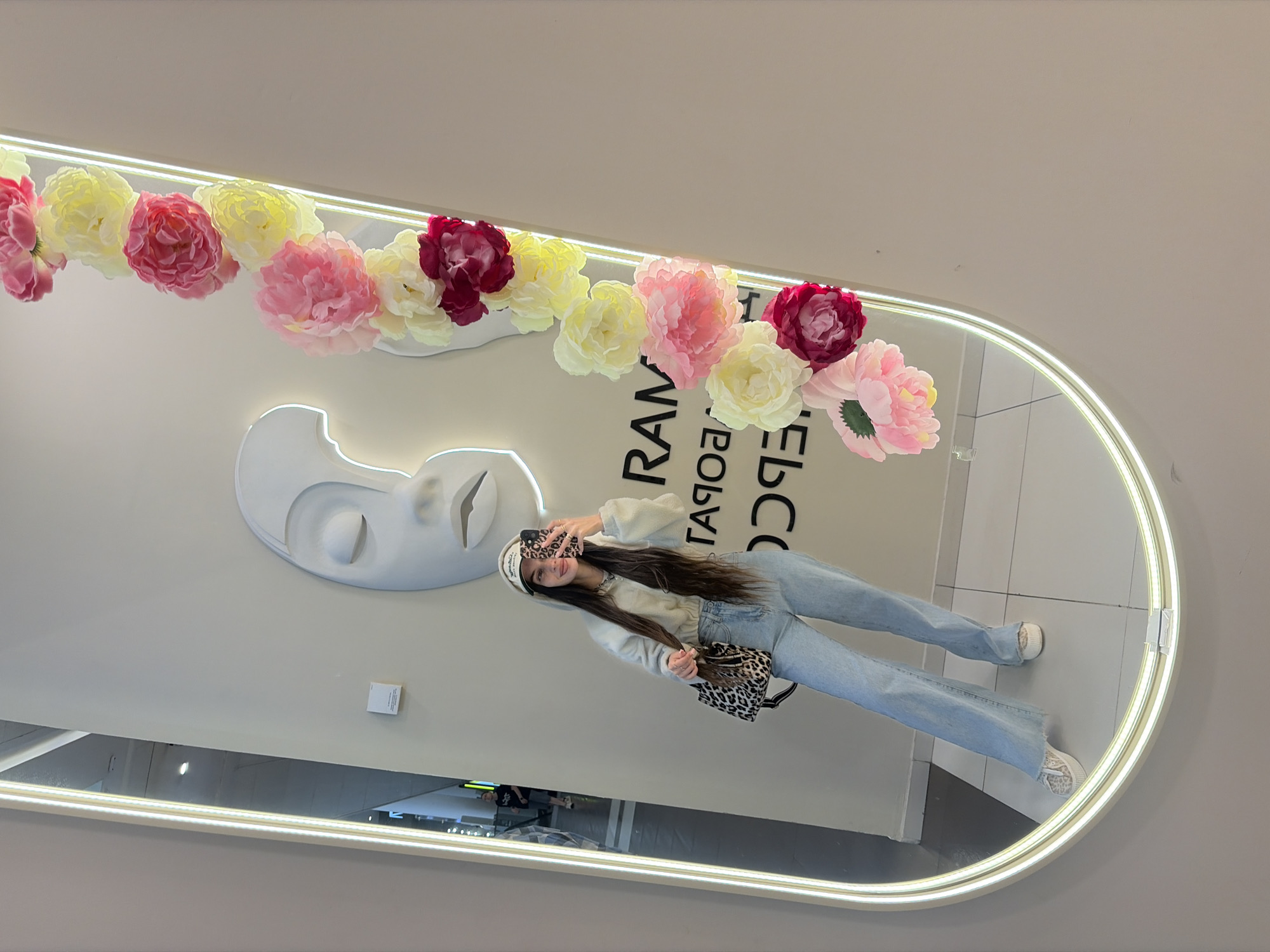 A young woman is taking a mirror selfie in what appears to be a retail store in Saint Petersburg, Russia. She is wearing a light-colored hooded sweatshirt, light blue denim flare jeans, a white and beige patterned baseball cap, and patterned sneakers. She is holding a smartphone with a leopard print case and a matching leopard print handbag on her left arm. Her long dark hair is visible, extending past her shoulders. The background features a large, oval mirror with a bright LED light strip around its perimeter. A white, abstract facial sculpture is mounted on the wall behind her, and a garland of artificial flowers in shades of pink, yellow, and burgundy cascades down the side of the mirror. Text is visible on the wall, appearing to be reversed in the mirror's reflection, possibly indicating store signage. The floor is tiled, and a blurred reflection of a person walking in the distance is visible in the mirror's left side.
