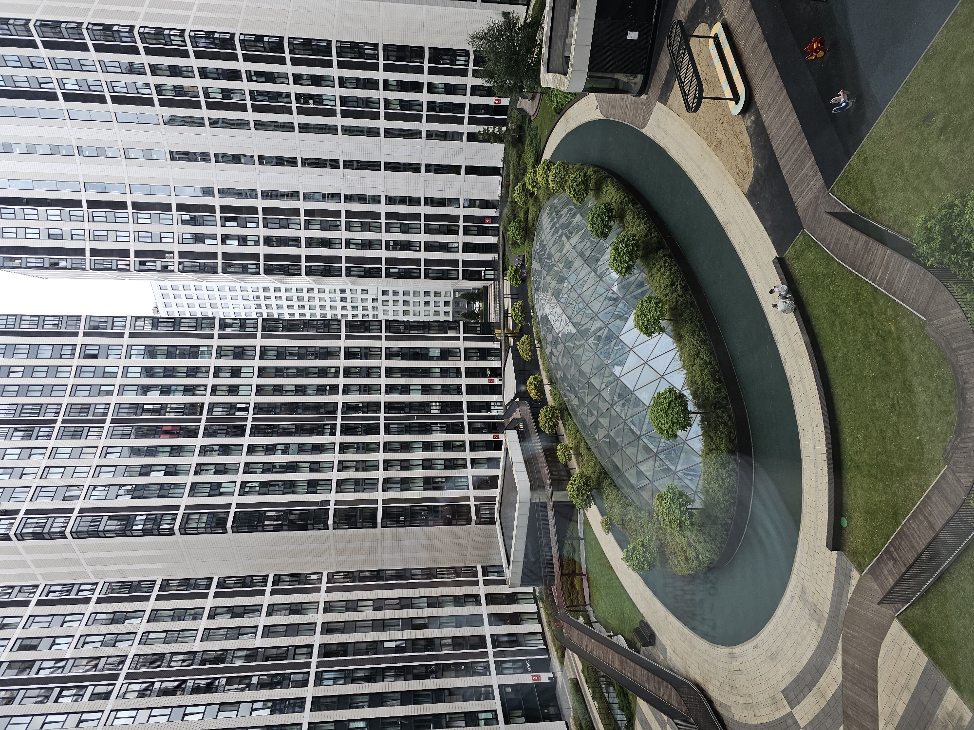An overhead shot captures a modern urban courtyard with towering residential buildings surrounding a central green space. The buildings, clad in light-colored panels and dark-framed windows, are arranged in a crescent shape. In the center of the courtyard is a distinctive geodesic dome structure made of glass, surrounded by manicured bushes and small trees. A dark, water-filled channel encircles the dome, bordered by a light-colored paved pathway. Wooden walkways and benches are interspersed throughout the courtyard, which also features a children's playground with two small rocking toys. Two individuals are seated on a bench, facing the dome. The overall scene depicts a well-designed, contemporary residential complex.