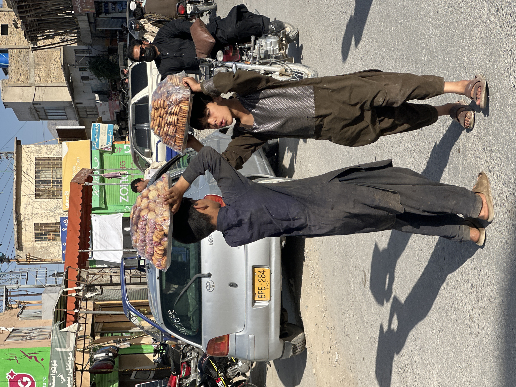 This daytime image captures a bustling street scene, likely in Sindh, Pakistan, given the visible license plate. The primary subjects are two young boys, seemingly street vendors, each balancing a large, clear plastic bag filled with cookies or biscuits on their heads. The boy in the foreground wears a dark, possibly black or dark blue, loose-fitting outfit and sandals, while the boy behind him wears a brown, similar-style outfit. Both have shadows trailing behind them on the sunlit asphalt, indicating a bright, clear day.
In the background, a silver Toyota Vitz car is parked, with its license plate "BPB-284 SINDH" clearly visible. Several motorcycles are also parked or being ridden. The street is lined with buildings featuring a mix of brick and concrete construction, and various storefronts with awnings and signage. One sign, partially obscured, displays "Mobile" and "Cash," along with "easypaisa" and "Zong 4G," suggesting mobile banking or communication services. Other signs show fragmented Urdu/Sindhi text, including "فوٹواست", "موبائل ریپی" (mobile repair), and "لا تسرع ذكر الله" (Do not rush the remembrance of Allah). An adult male in a dark shirt and face mask is visible near a white car in the mid-ground. The scene portrays a slice of daily urban life, with the boys engaged in their work amidst the street's activity.